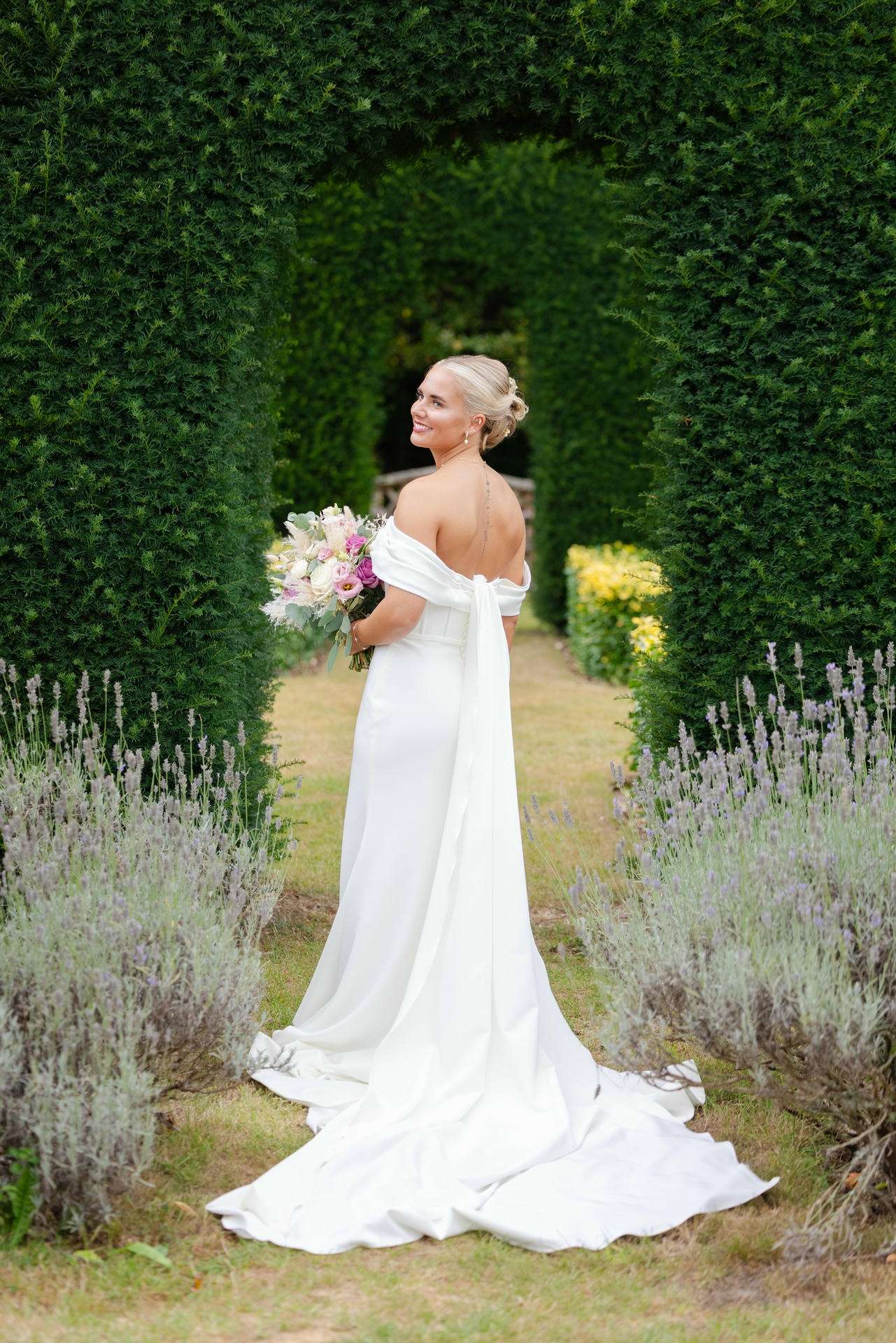 A bride in a white off-the-shoulder wedding gown with a long train stands in a hedge-arch garden, holding a pastel bouquet among lavender plants at The Gardens Wedding venue