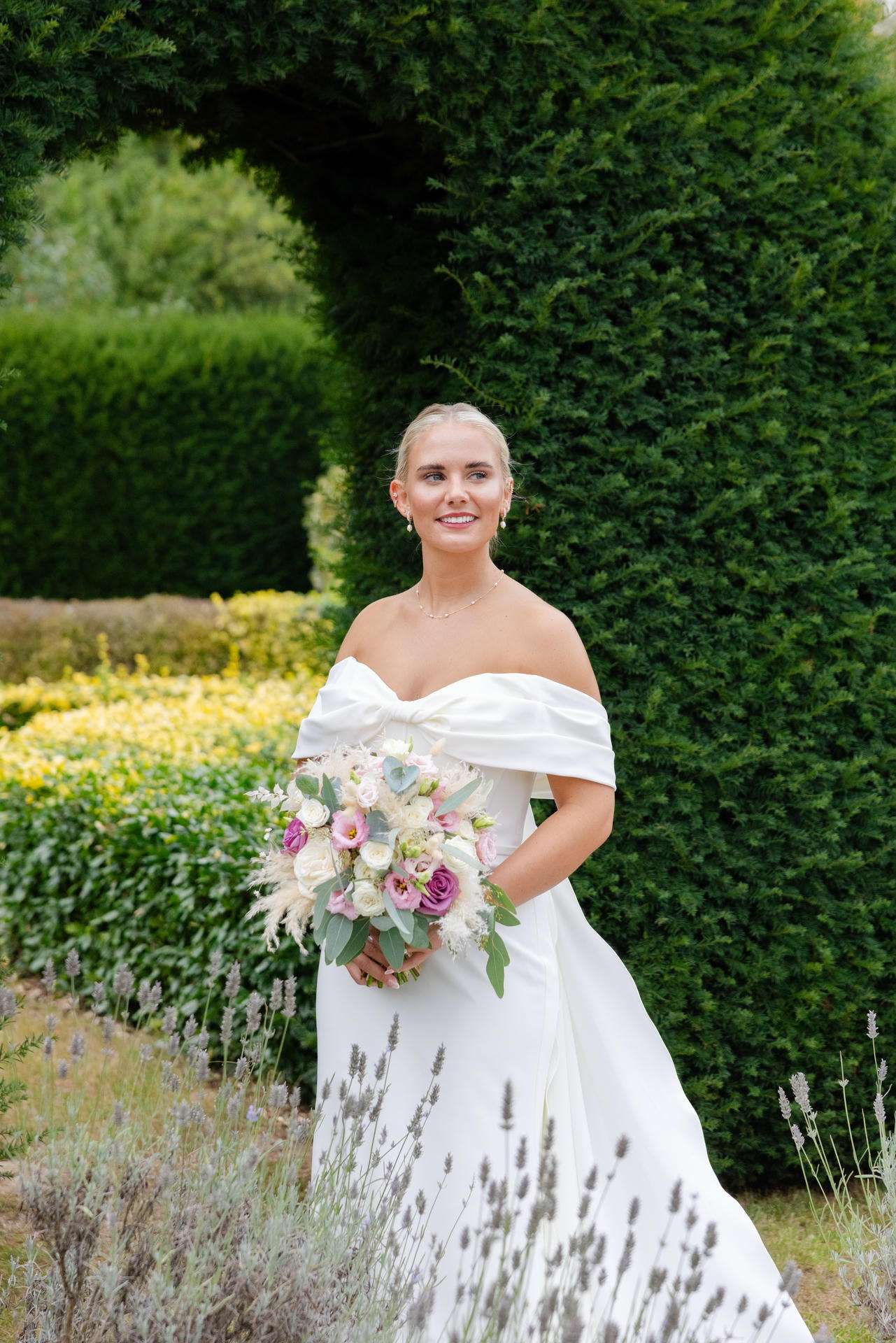 A bride in an off-the-shoulder white wedding gown holds a pastel bouquet in a lush garden at The Gardens Wedding venue