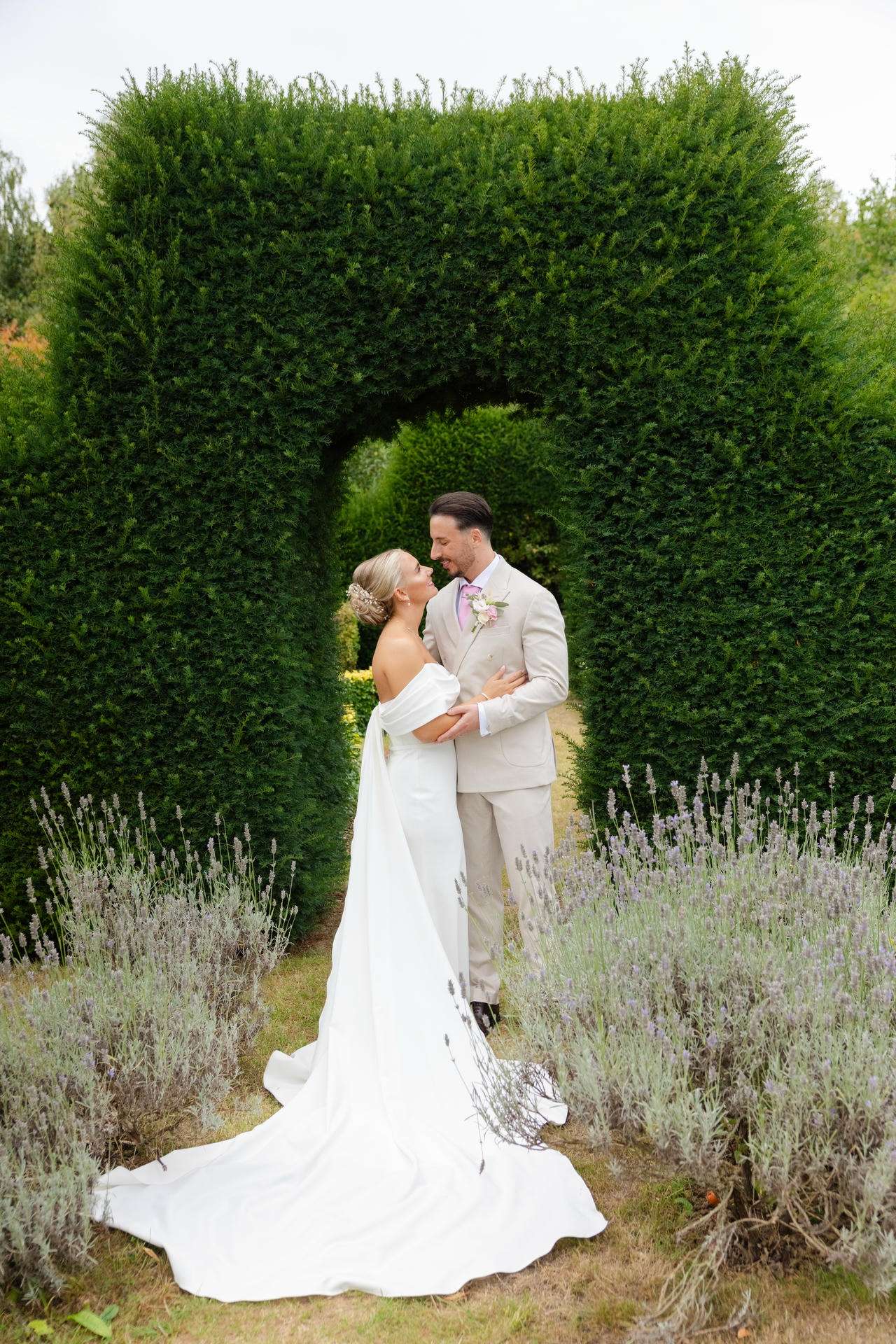 Bride and groom embrace under a tall, square hedge arch in a lavender-filled garden.