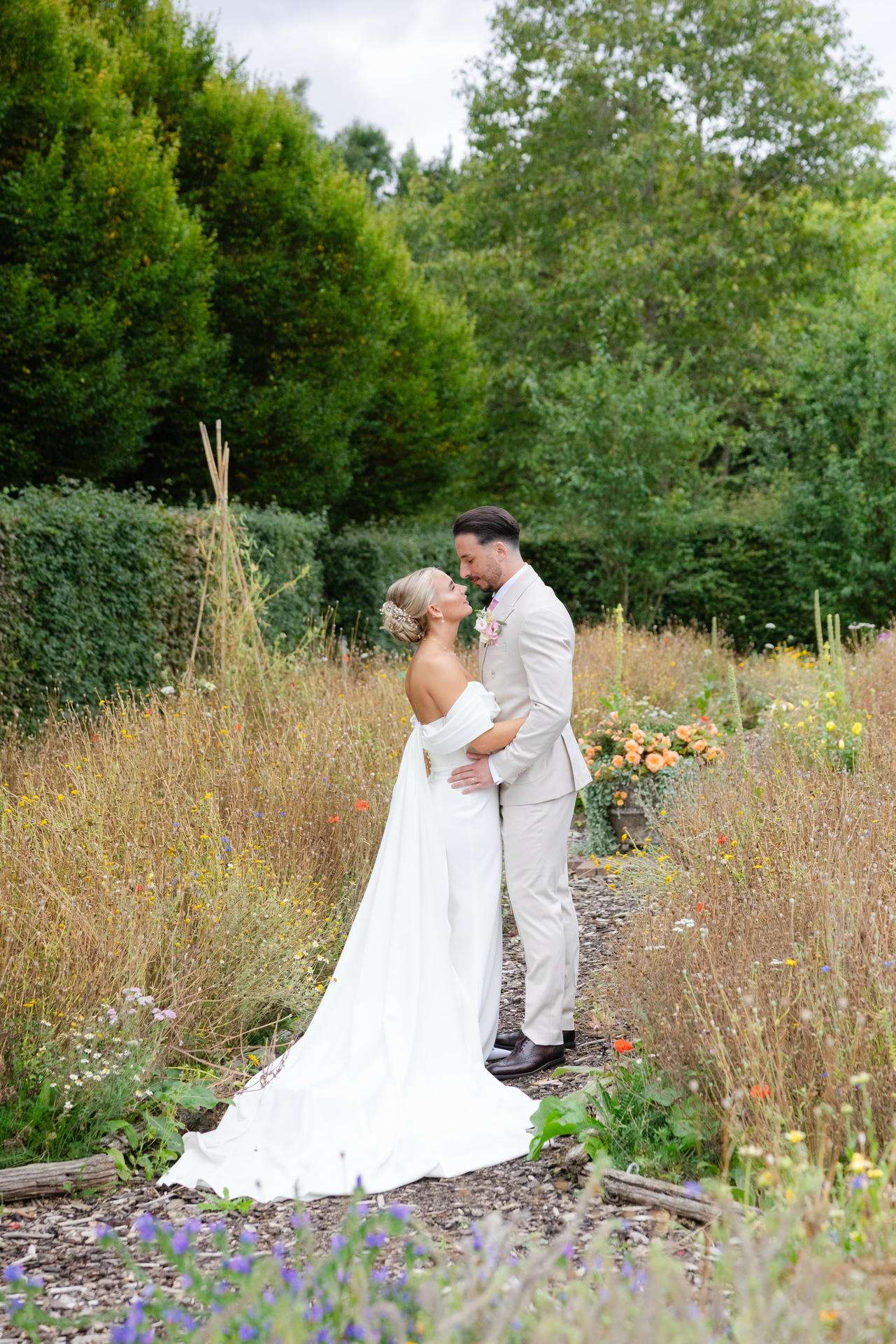 A bride in a white wedding gown with a long train and a groom in a light suit share a hug on a stone path amid a lush garden of wildflowers.