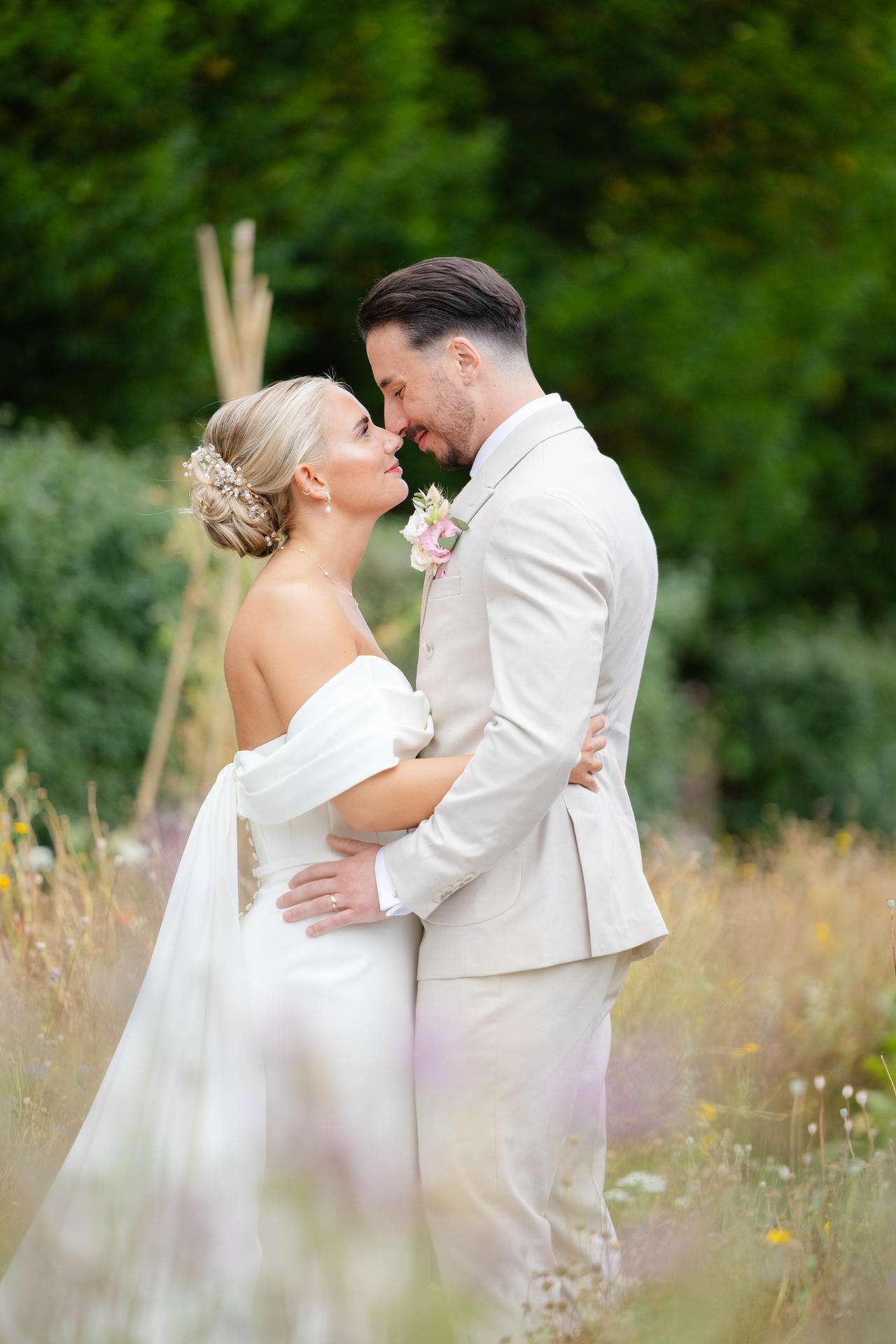 Bride and groom embrace in a meadow, the bride in an off-shoulder white wedding gown and the groom in a light beige suit at The Gardens Wedding venue