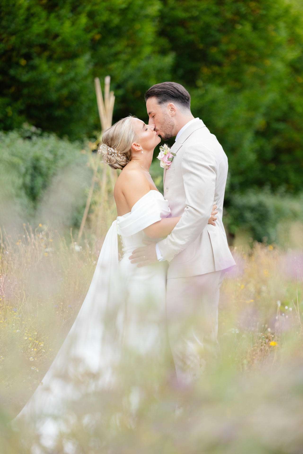 A bride in an off-shoulder white wedding dress and a groom in a light beige suit kiss in a meadow filled with wildflowers.
