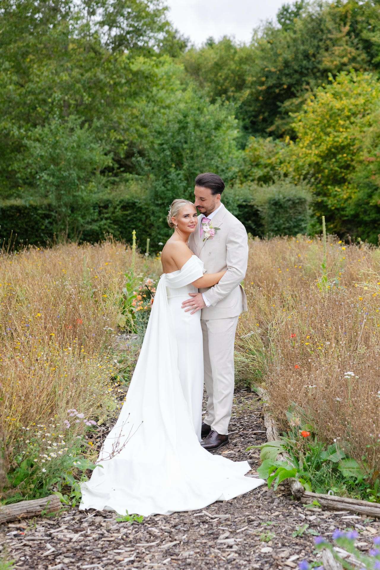A bride in a white off-shoulder wedding dress hugs a groom in a light beige suit in a meadow of tall wildflowers and greenery at The Gardens Wedding venue