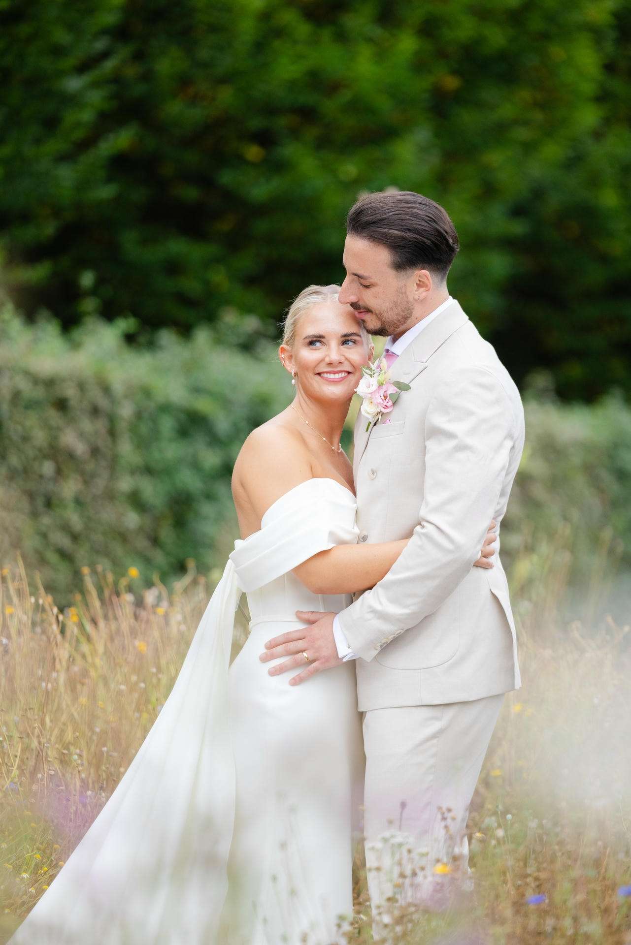 A bride in an off-shoulder white wedding dress and a groom in a light beige suit embrace in a meadow of wildflowers as he kisses her temple at The Gardens Wedding venue