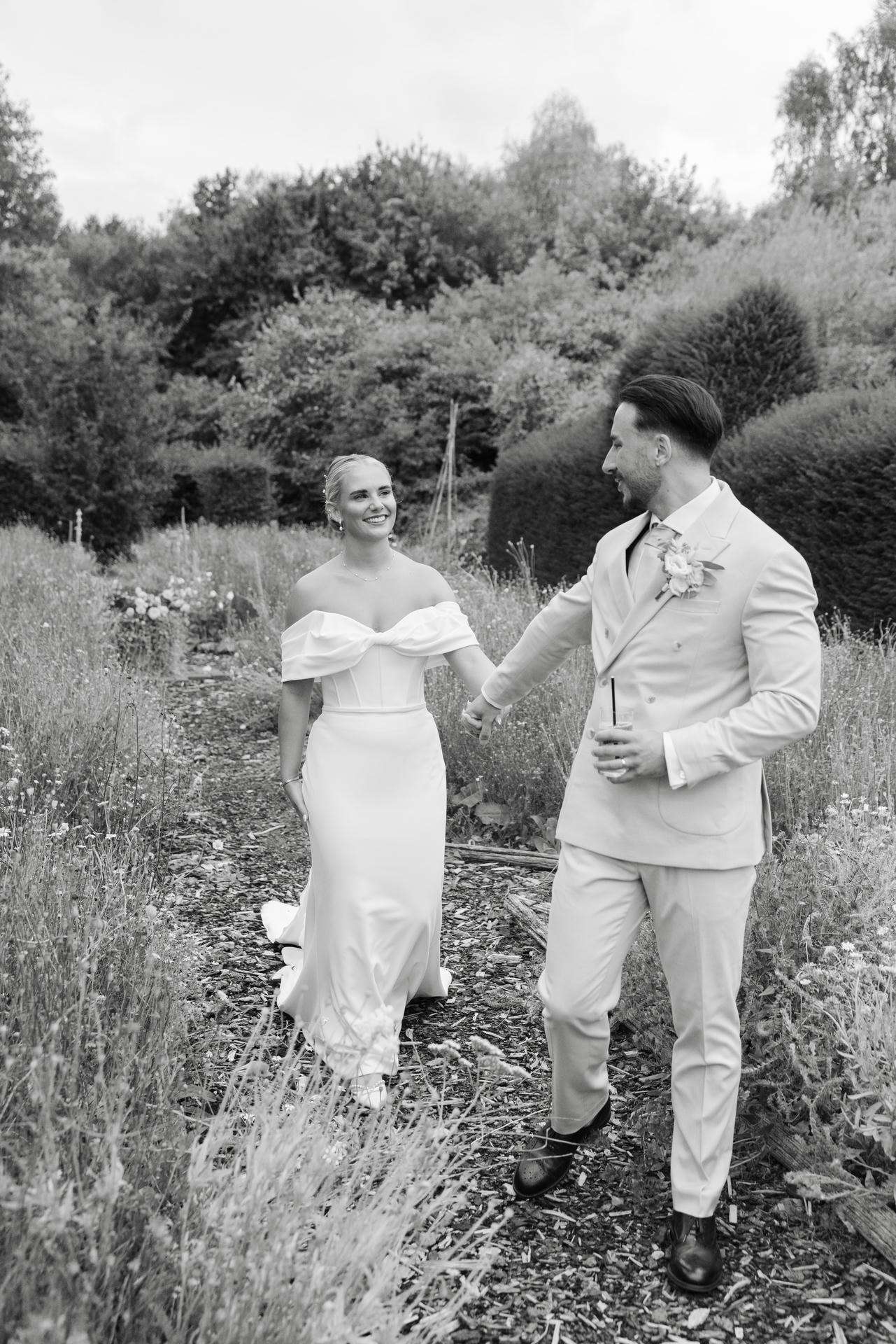 A smiling bride in an off-shoulder white wedding gown and a groom in a light suit walk hand in hand along a garden path at The Gardens Wedding venue