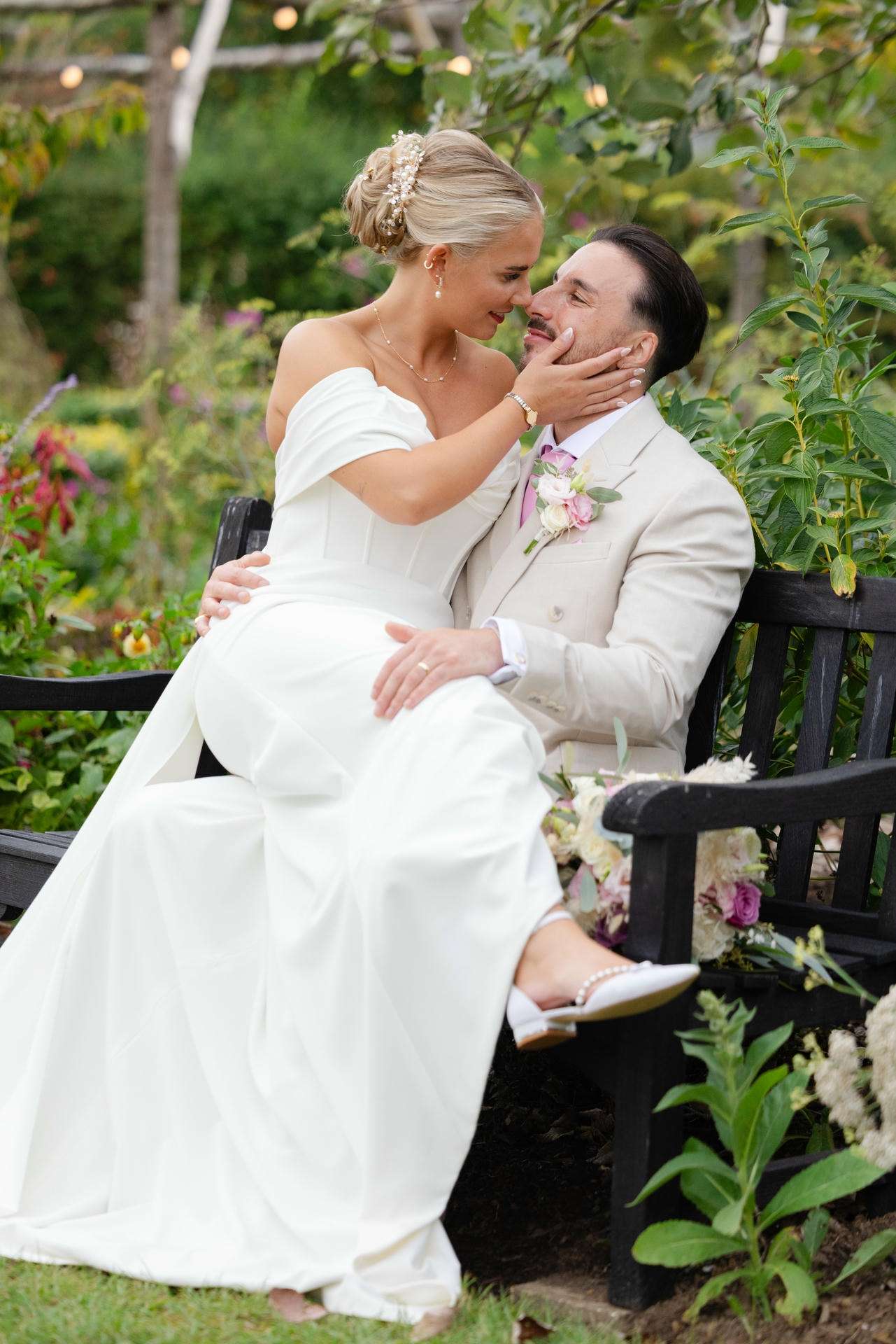 Bride in a white off-shoulder wedding dress sits on her groom's lap on a garden bench, tenderly cupping his face at The Gardens Wedding venue