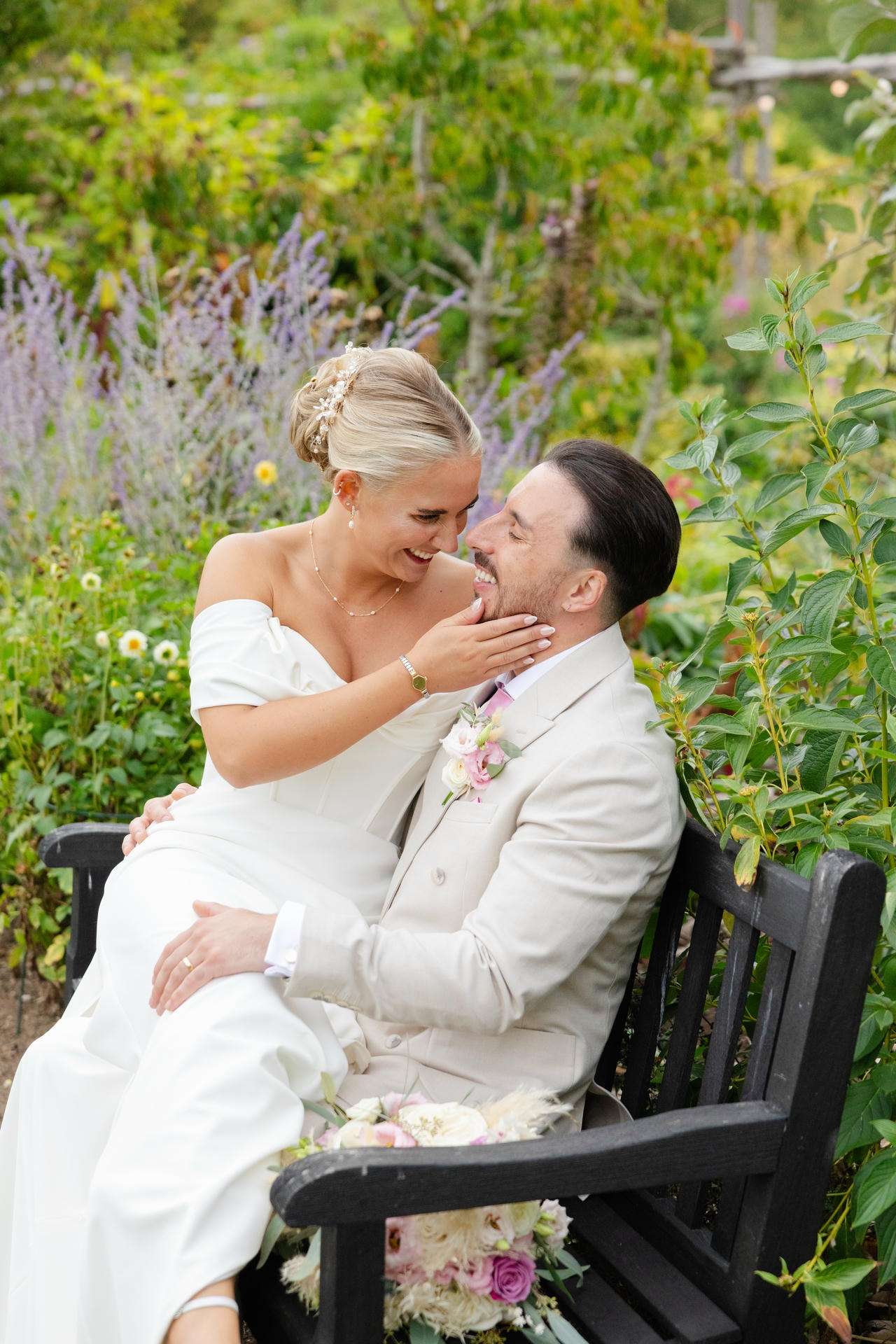 A bride in a white off-shoulder gown and a groom in a light suit sit close on a garden bench, smiling and touching faces amid blooming flowers at The Gardens Wedding venue