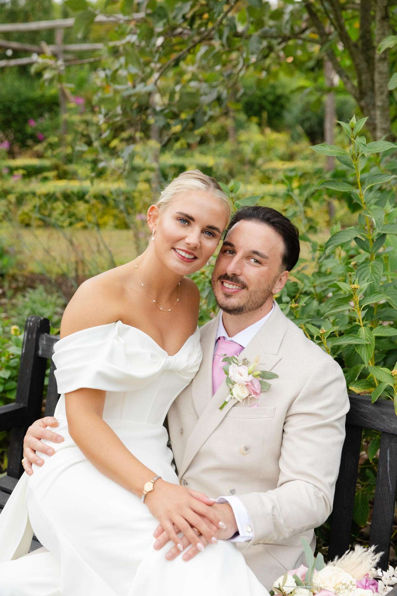A bride in an off-the-shoulder white gown and a groom in a light beige suit sit closely on a bench in a lush garden, smiling at the camera at The Gardens Wedding venue