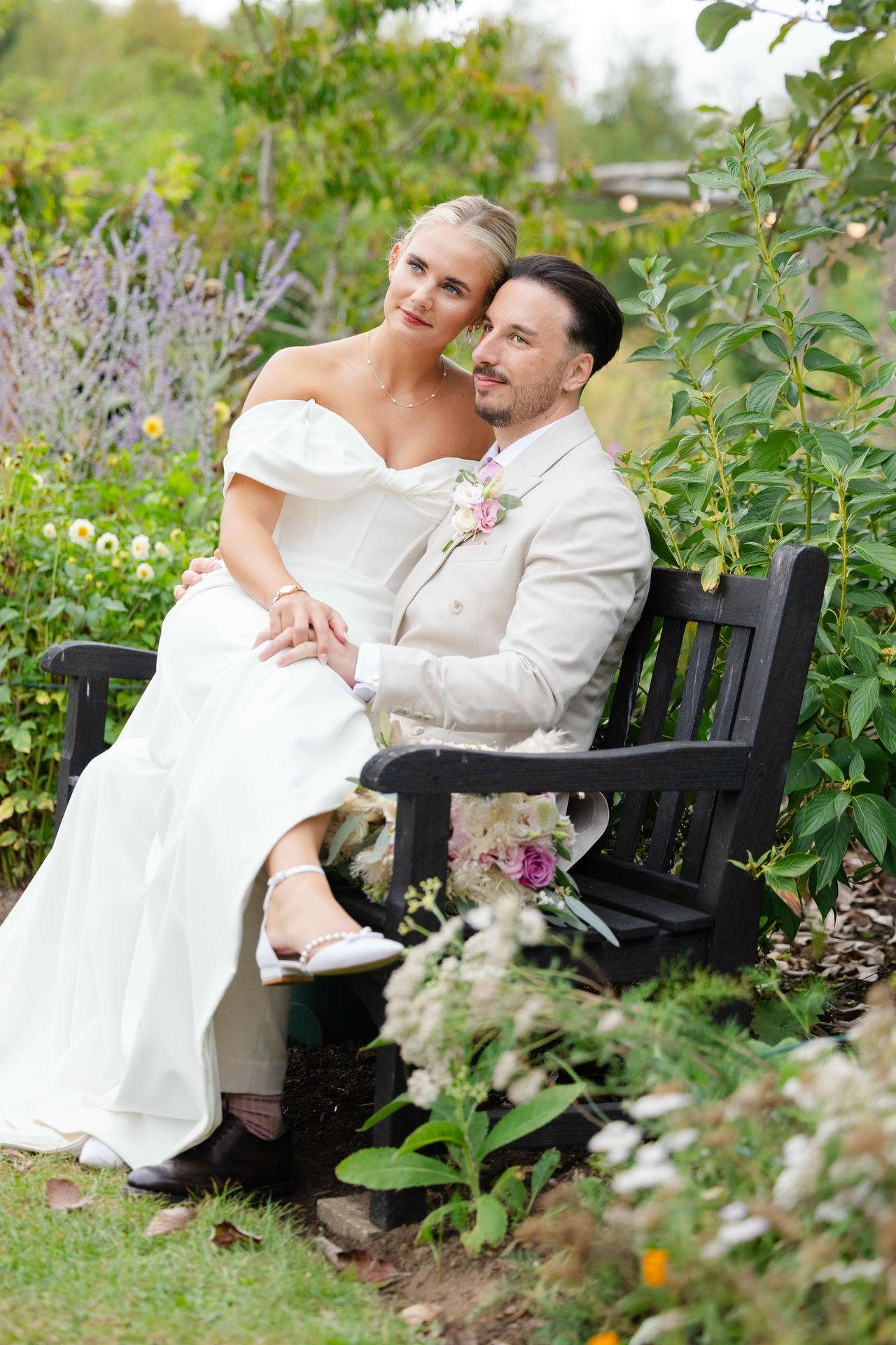 A bride and groom sit close together on a black bench in a lush garden, the bride in an off-the-shoulder white gown and the groom in a light beige suit at The Gardens Wedding venue