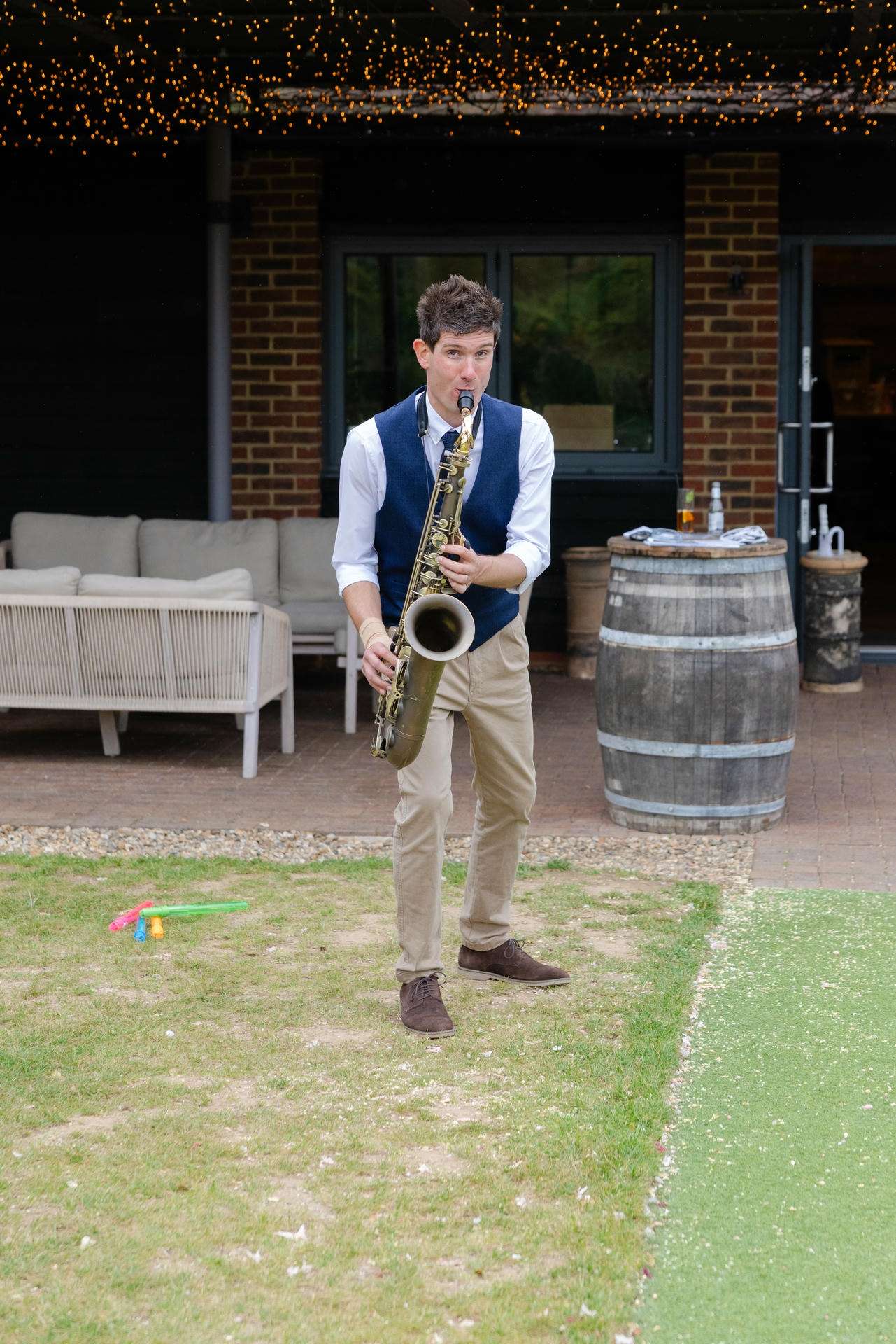 A man in a blue vest and beige pants plays a saxophone outdoors at a yard party, under string lights.
