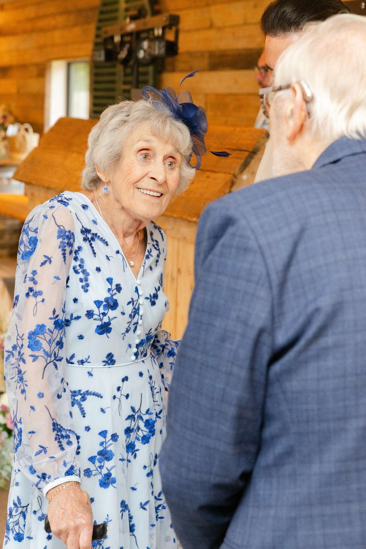 Smiling elderly woman in a light blue floral dress with a blue fascinator talks with a man in a blue suit.
