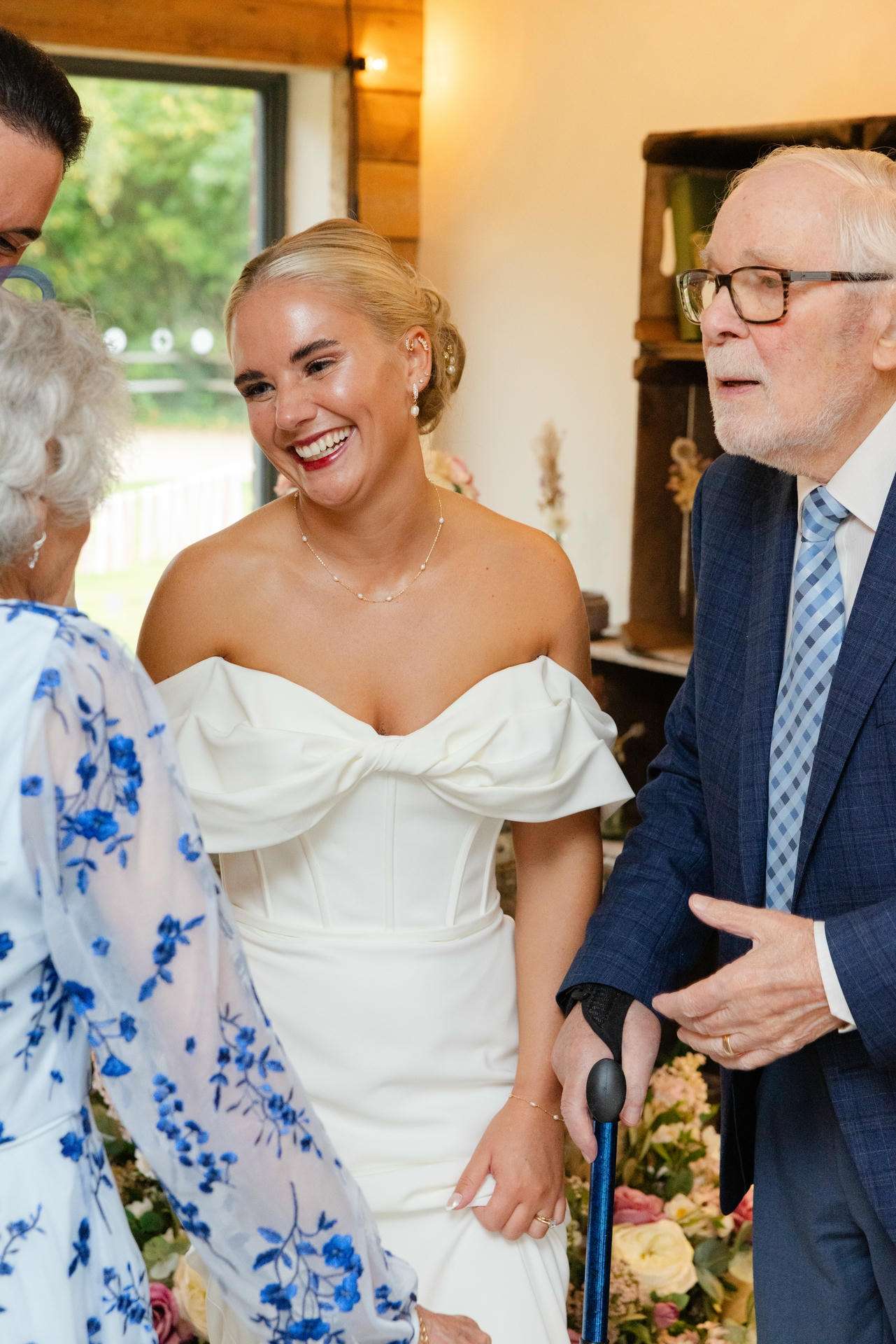 Smiling bride in an off-the-shoulder white wedding dress chats with an elderly man in a suit indoors.