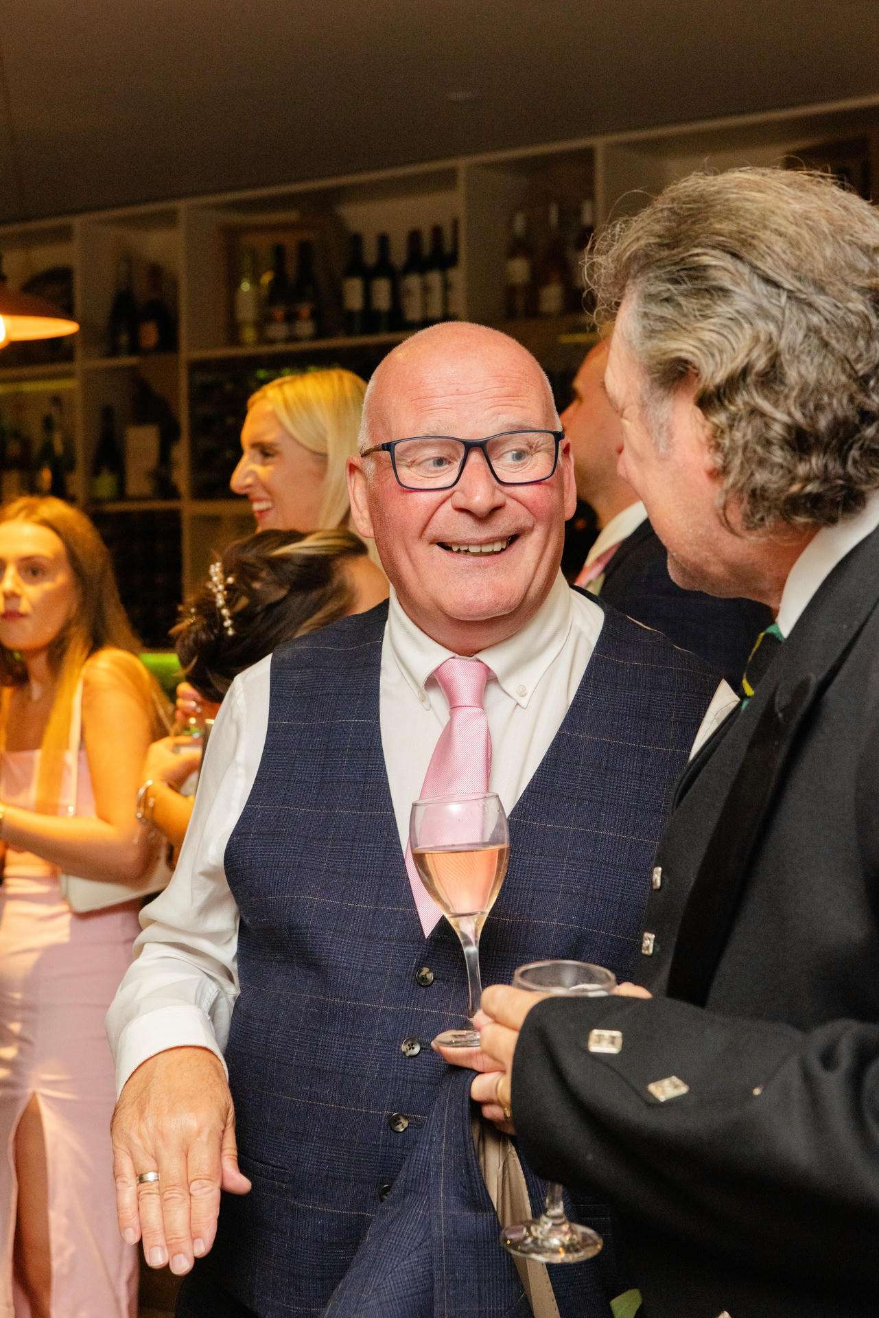 Smiling bald man in a blue plaid waistcoat and pink tie holds a glass of rosé while chatting with another man at a social event.