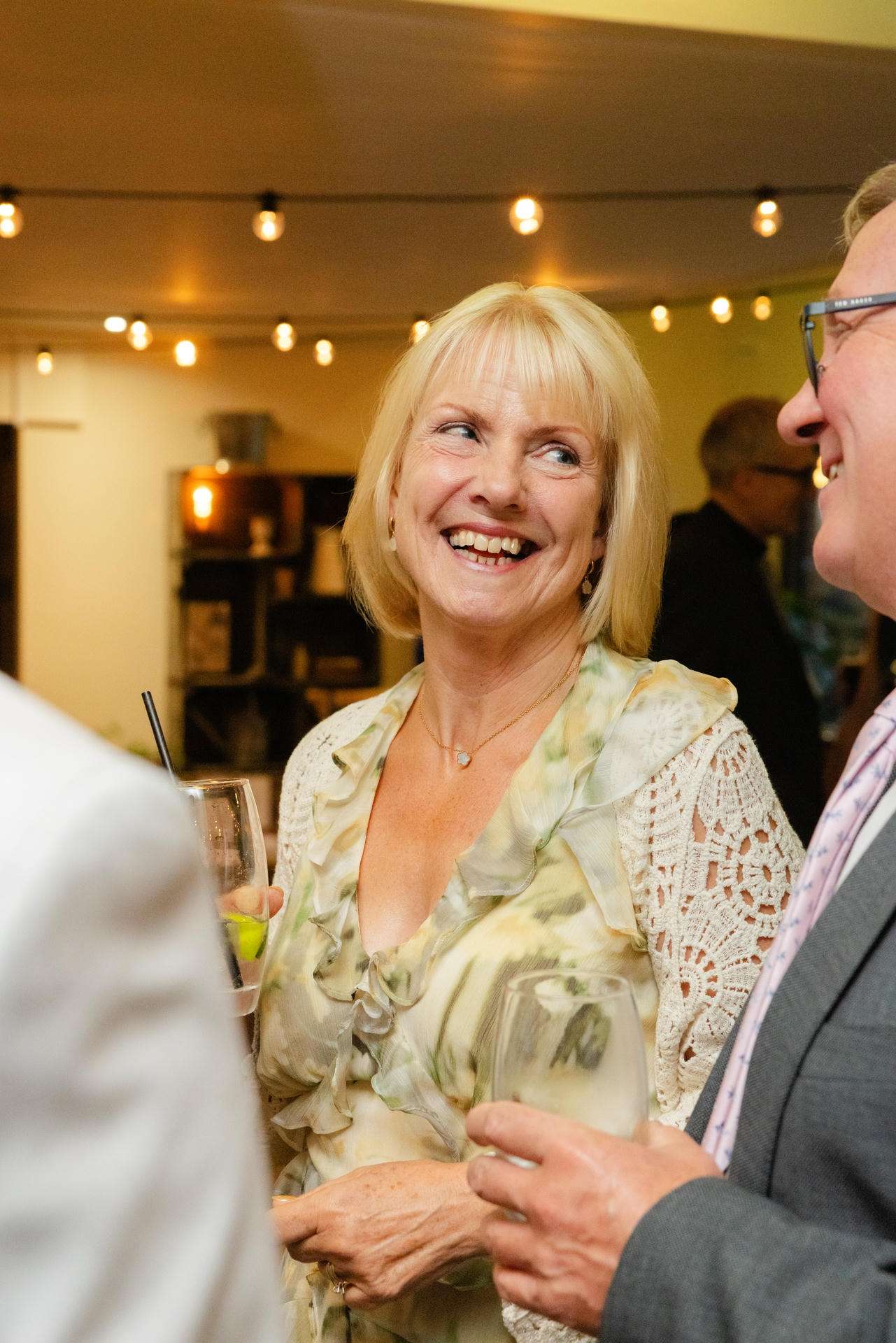 Smiling blonde woman in a light floral blouse and lace cardigan holding a glass while chatting at a social gathering.