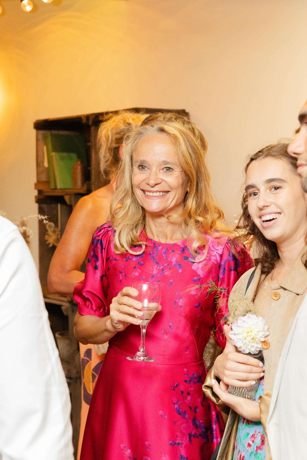 Smiling woman in a bright pink floral dress holds a champagne glass at an indoor party, standing beside a younger woman with a flower accessory.