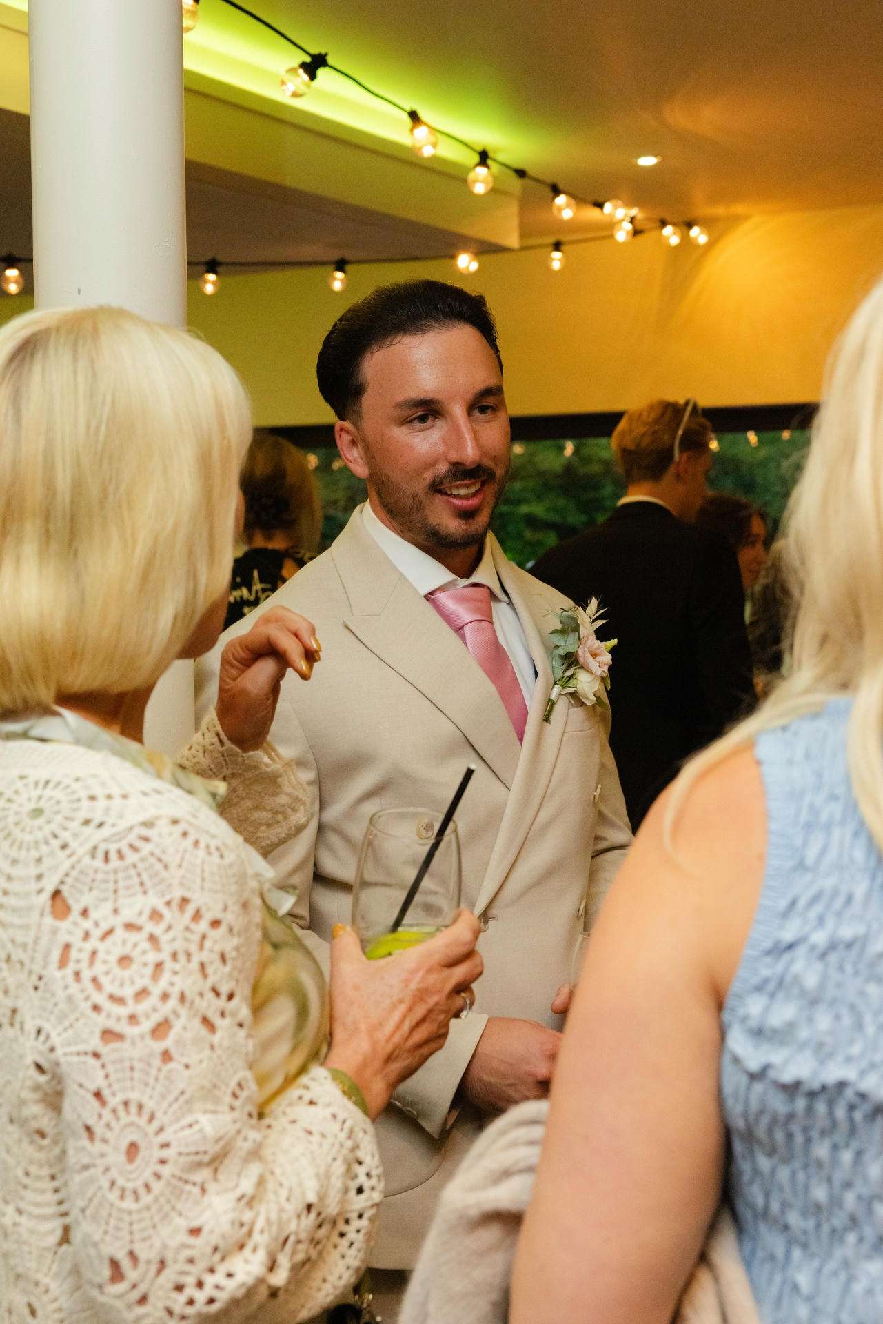 A man in a light beige suit with a pink tie and floral boutonniere talks with guests at a wedding reception under string lights.