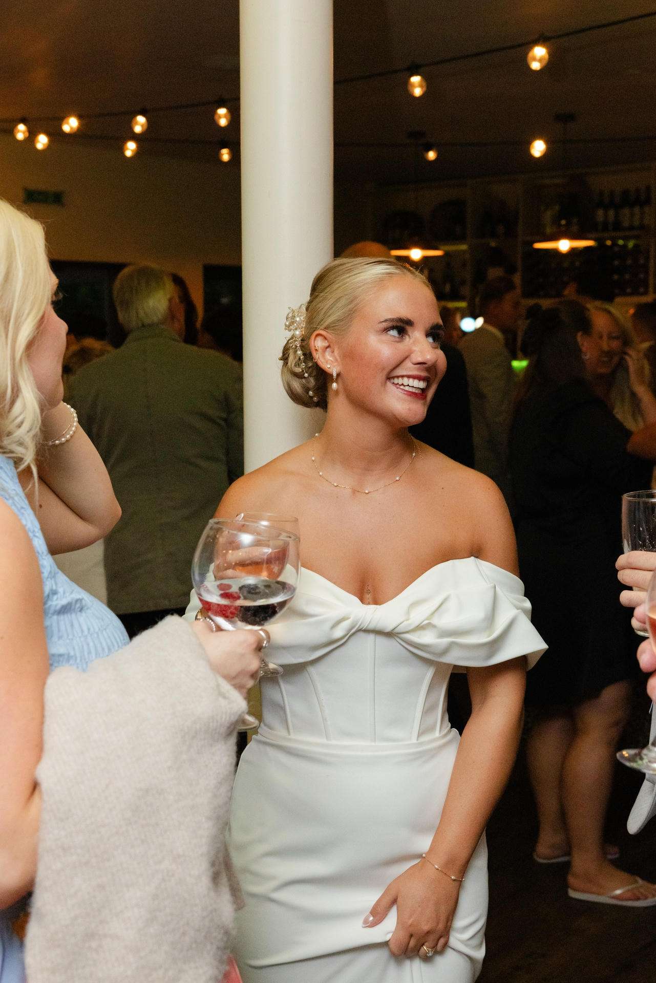 Smiling blonde woman in a white off-the-shoulder dress with an elegant updo at a reception.