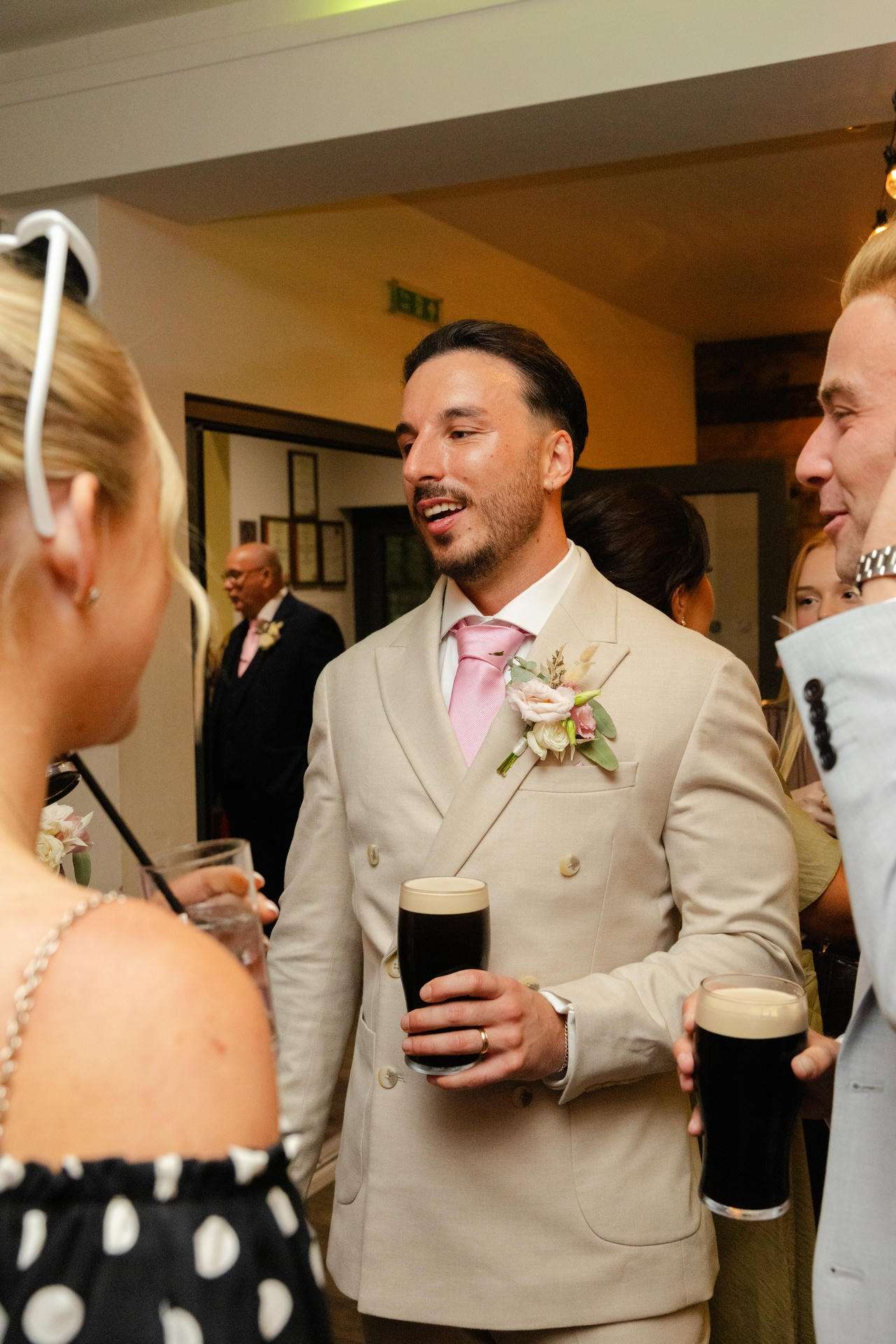 A man in a light beige suit with a pink tie and boutonniere holds a pint of dark beer while chatting with guests at a wedding reception.