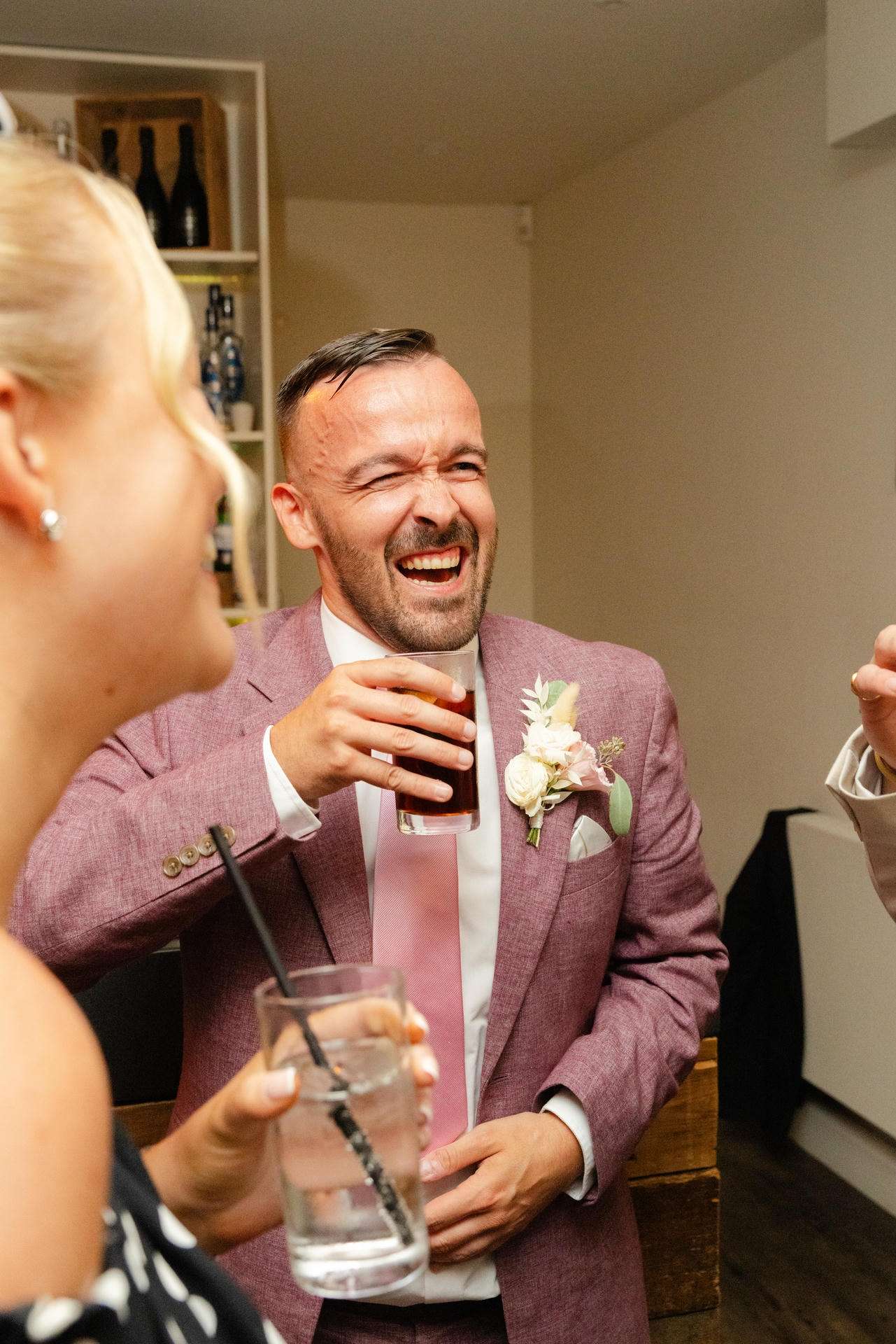 Smiling man in a pink suit with a boutonniere raises a glass of dark beverage at a party, with a woman in the foreground holding a drink.