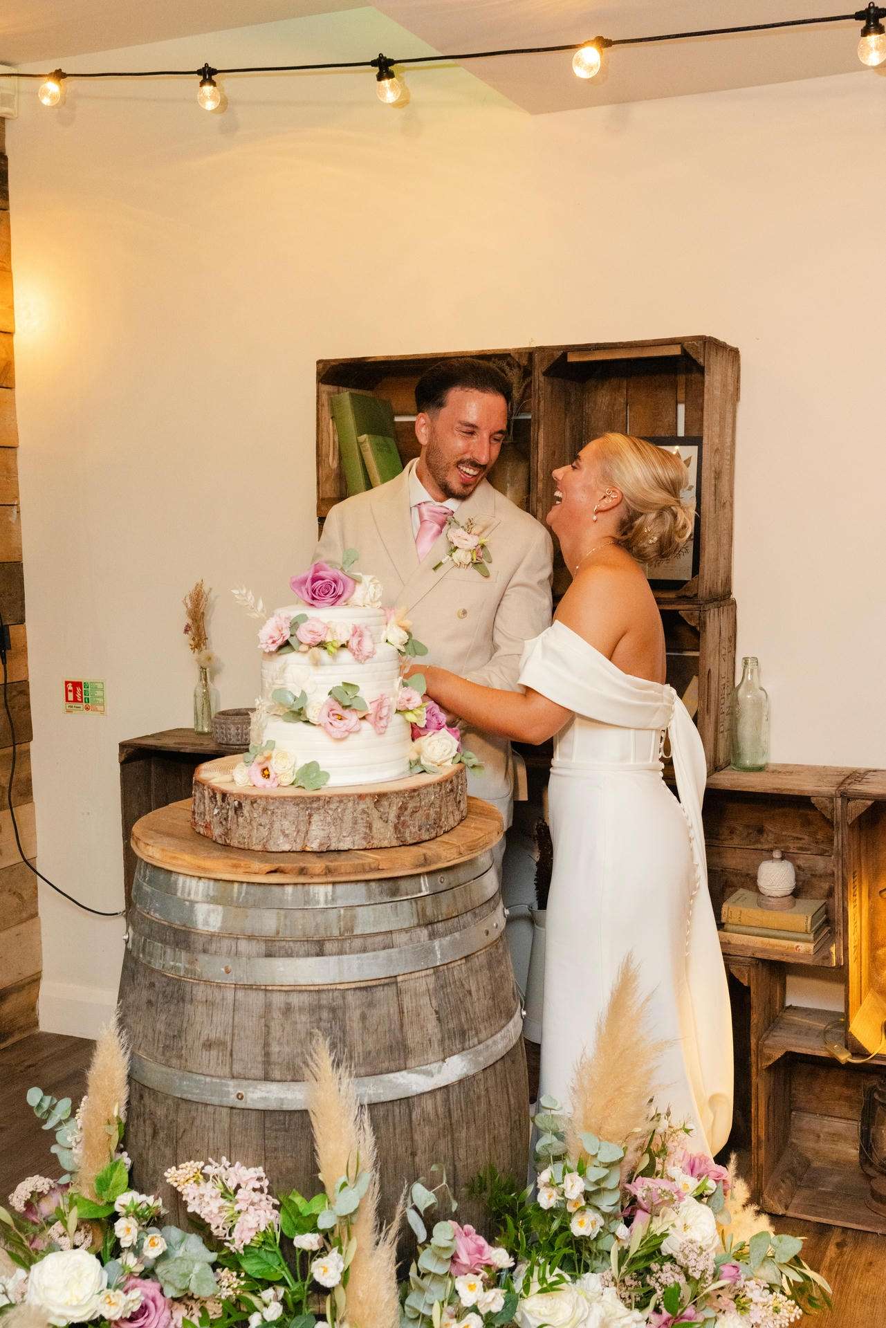 A bride and groom cut a three-tier wedding cake decorated with pink and white flowers on a wooden barrel in a rustic room.