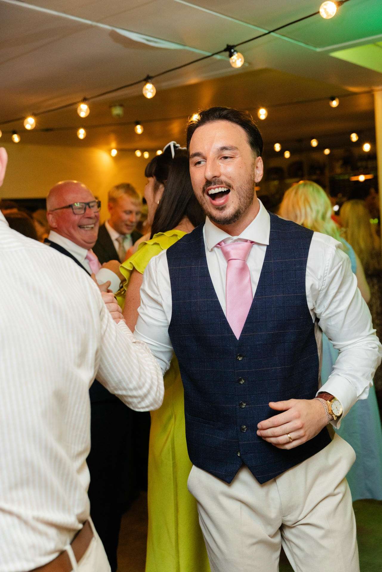 A man in a navy vest, white shirt, and pink tie dances at a lively indoor party with string lights and smiling guests in the background.