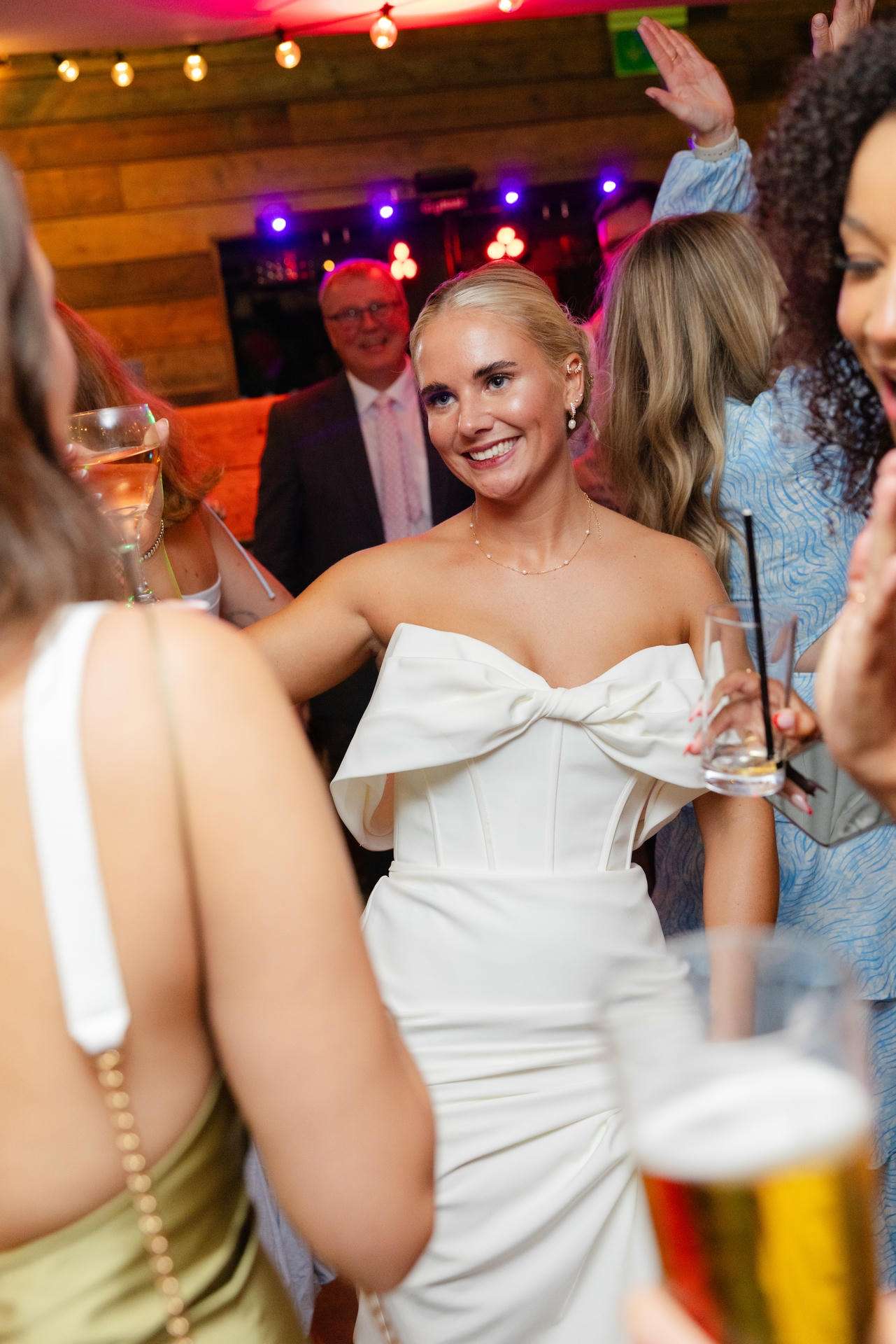 Smiling blonde woman in a white off-shoulder dress with a bow, holding a drink at a lively party.