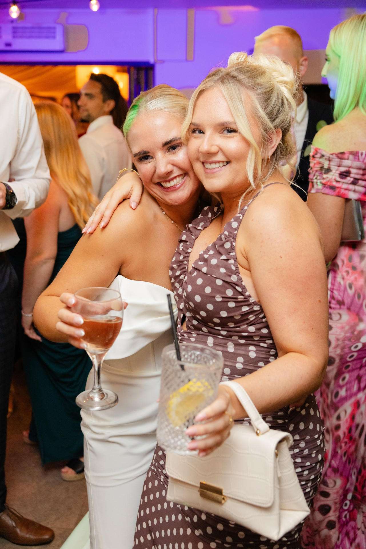 Two smiling women at a party hug for a photo, one in a white strapless dress holding a wine glass and the other in a brown polka-dot dress carrying a white purse.