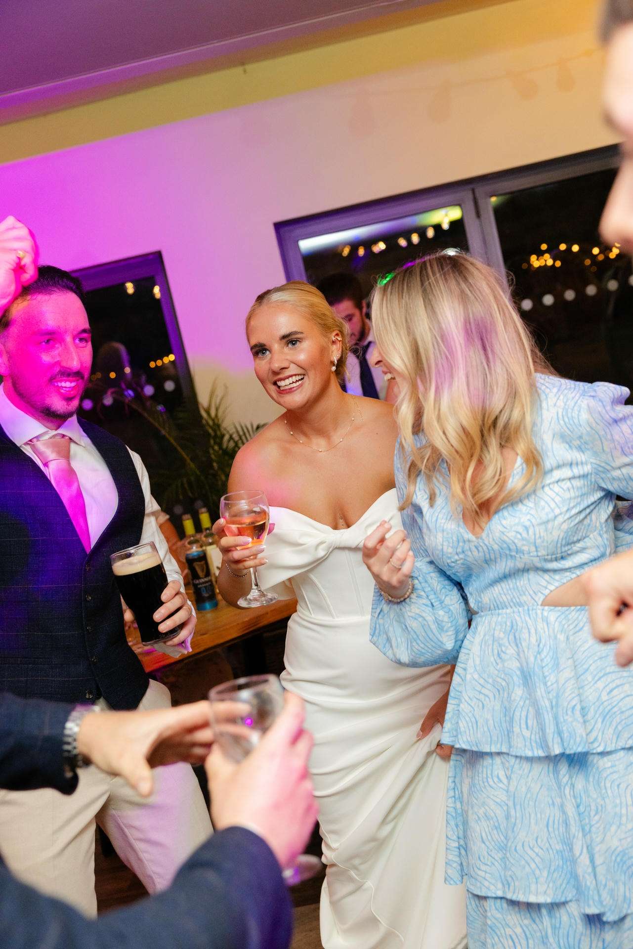 Bride in a white strapless dress laughs with friends at a party, holding a wine glass under colorful lighting.