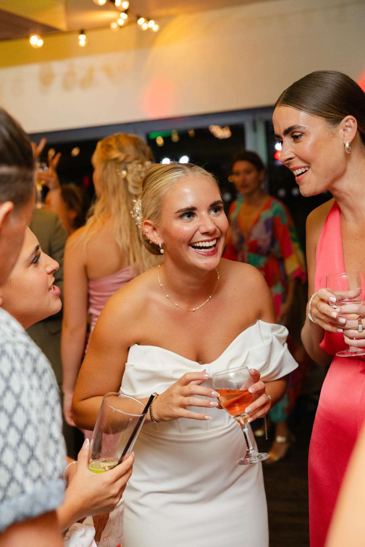 A smiling woman in a white off-shoulder dress chats with friends at a party, holding a glass of pink cocktail.