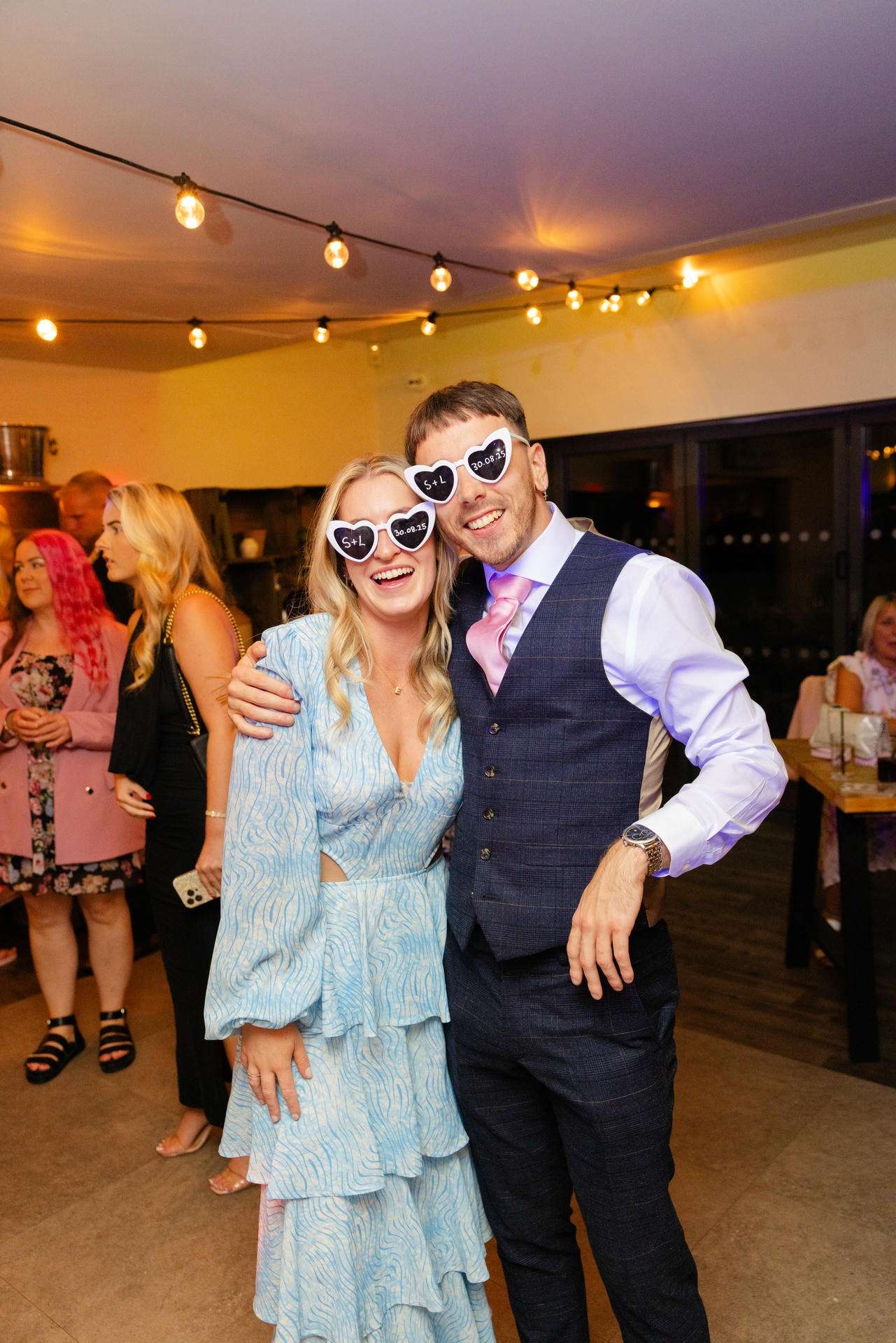Smiling couple posing at a party under string lights, with the woman in a light blue ruffled dress and heart-shaped sunglasses and the man in a navy vest with a pink tie.