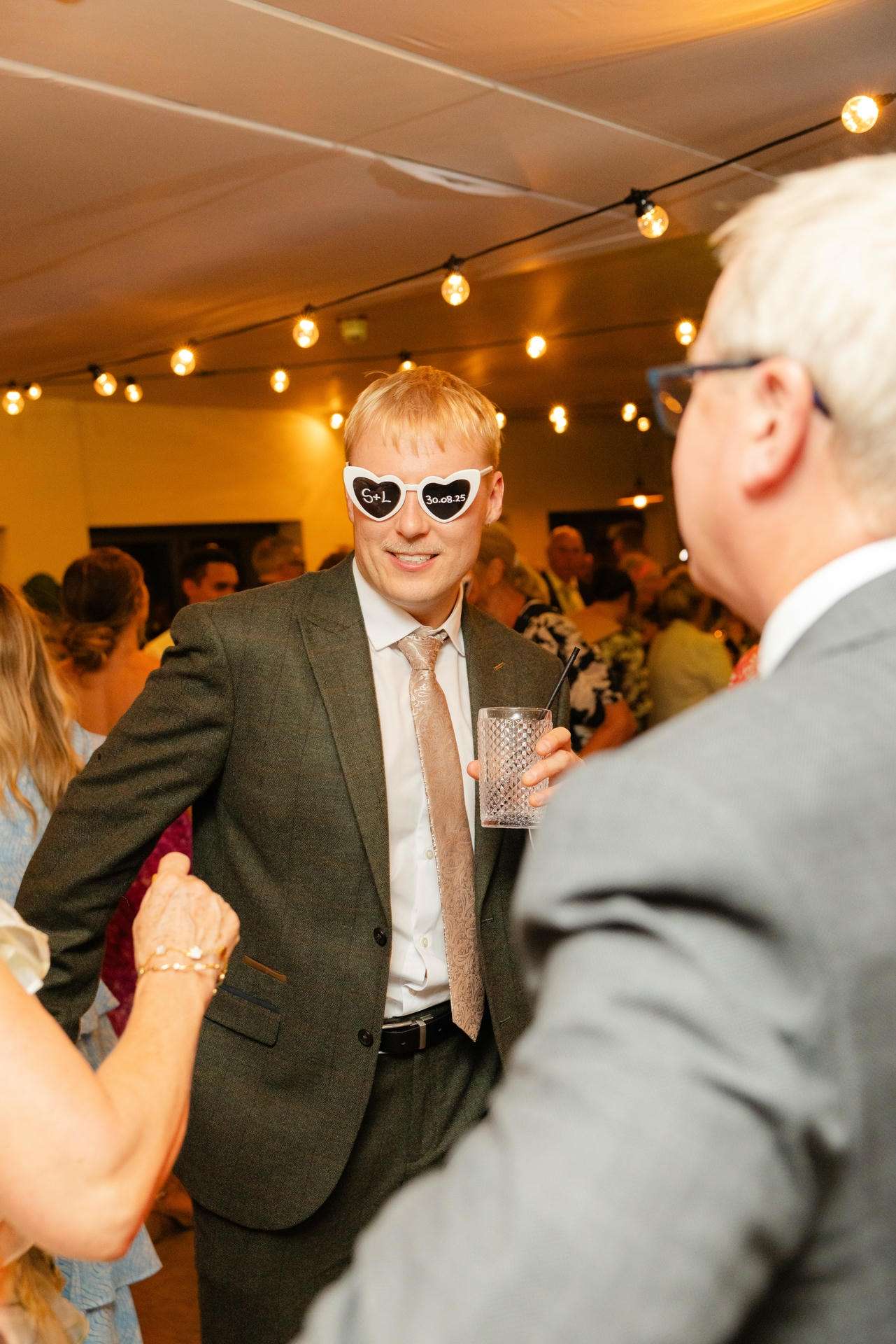 A man in a gray suit dances at a wedding reception, wearing heart-shaped sunglasses and holding a drink.