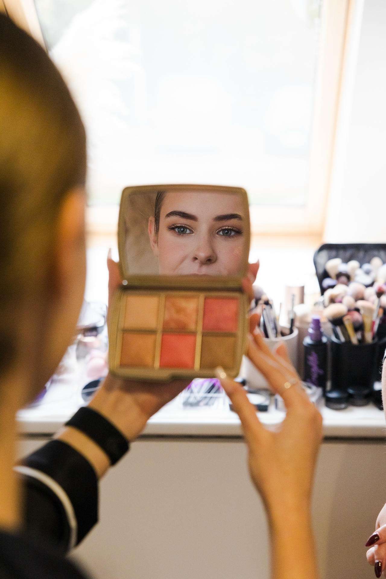 A makeup palette with six shades is held in front of a mirror showing a woman's face.