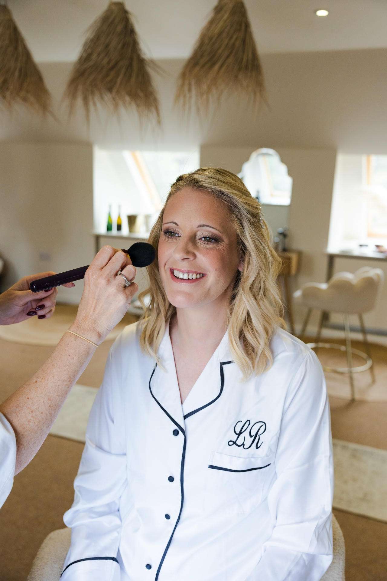 Smiling blonde woman in a white robe sits as a makeup artist applies makeup with a brush in a bright, modern salon.