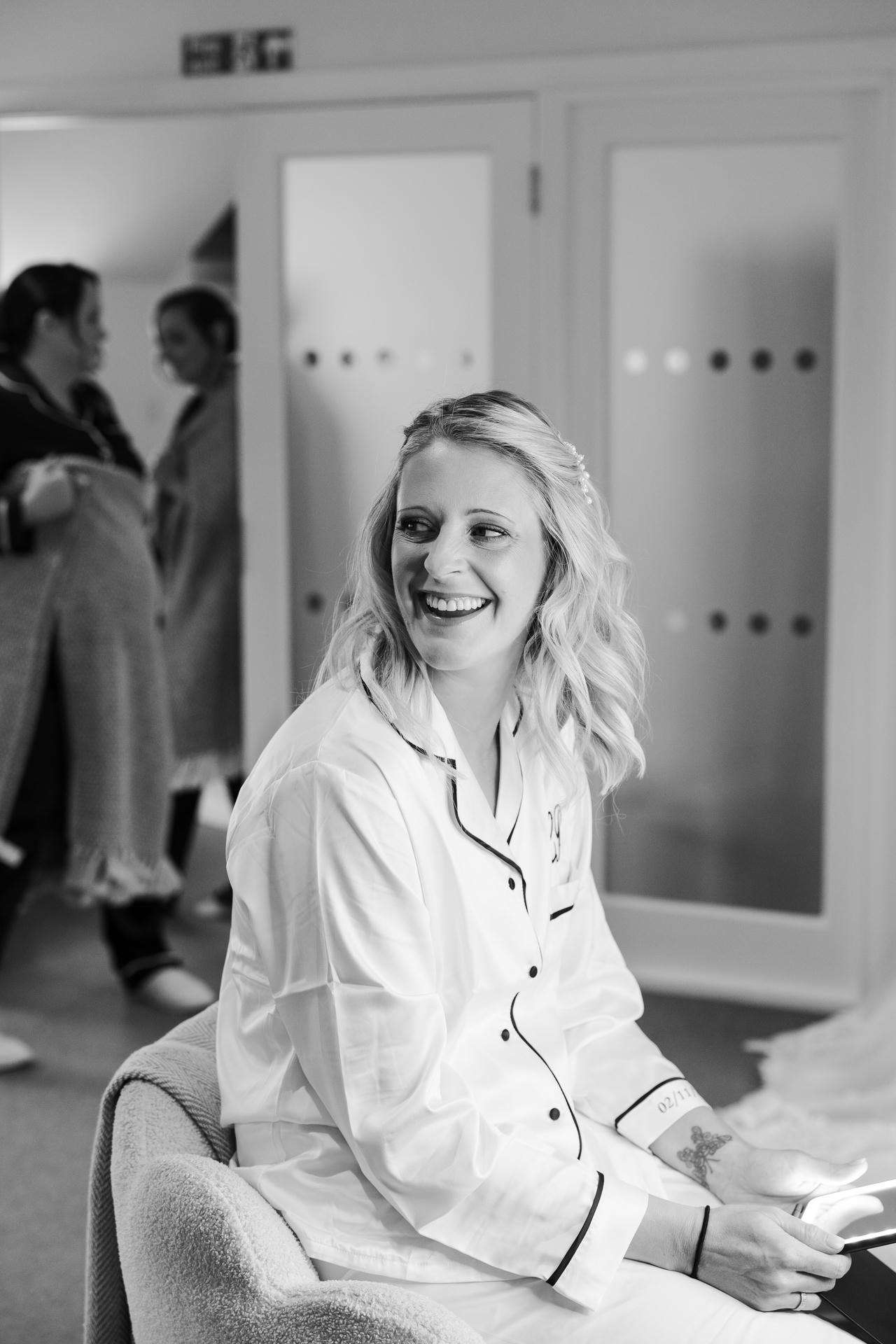 Smiling woman in a white pajama top sits on a chair in a room, with blurred people in the background.