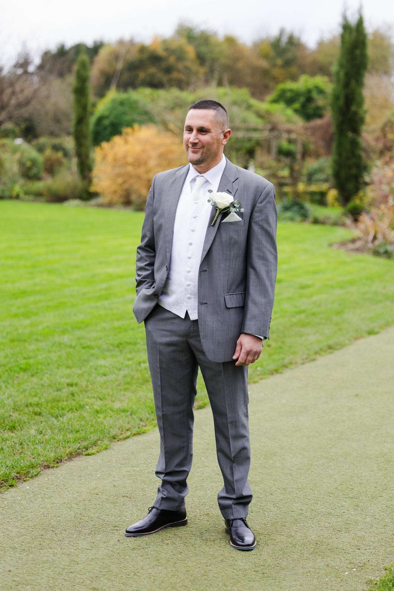 A man in a gray suit with a white waistcoat and boutonniere stands on a path in a lush garden.