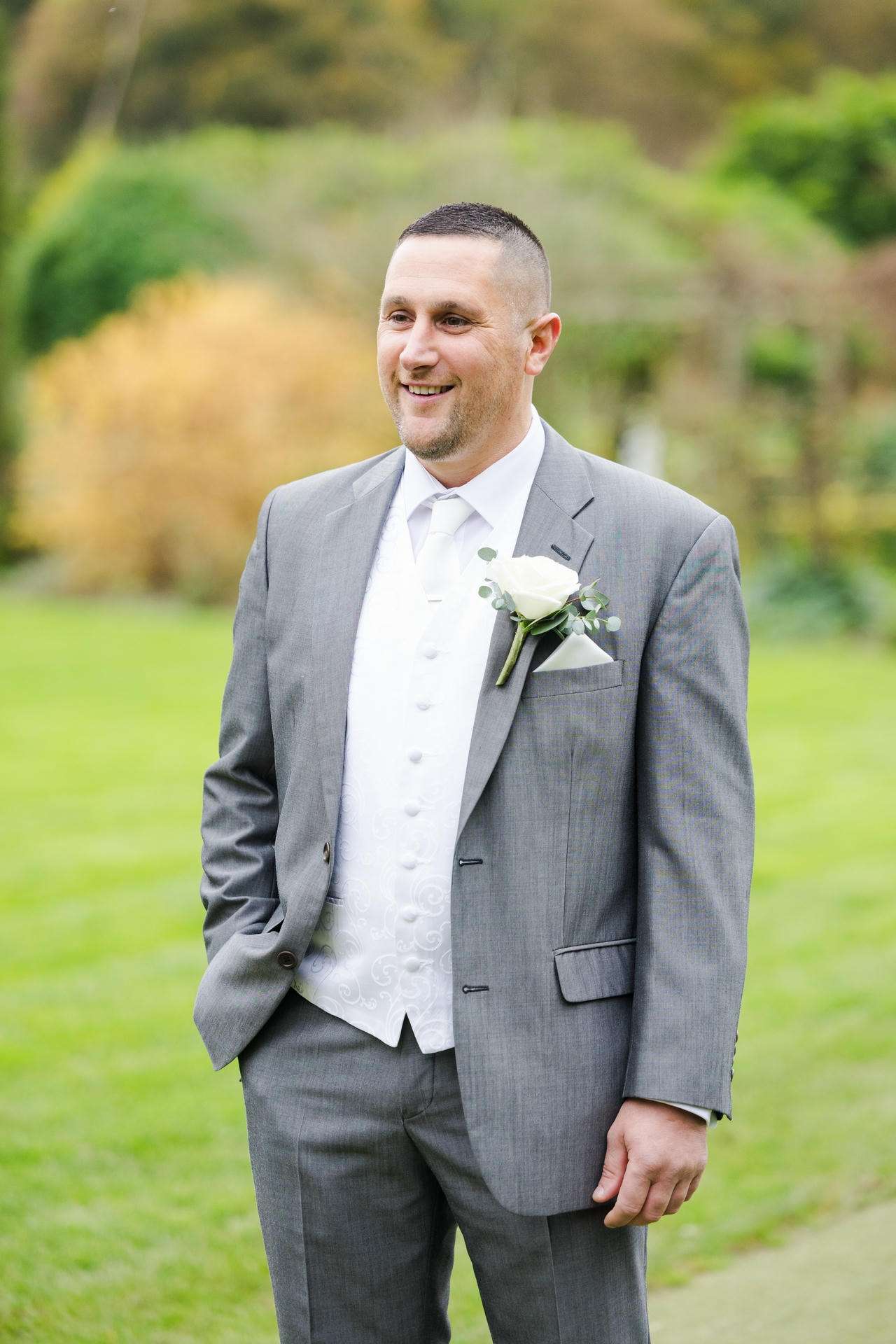 Man in a gray suit with a white waistcoat and boutonniere stands on a grassy lawn, smiling with a softly blurred garden background.