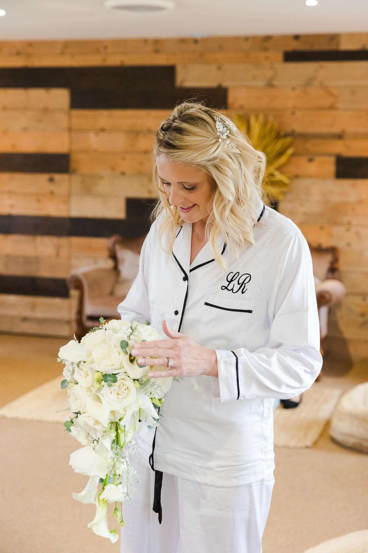 Blonde woman in a white robe with black piping, holding a cascading white bouquet indoors.