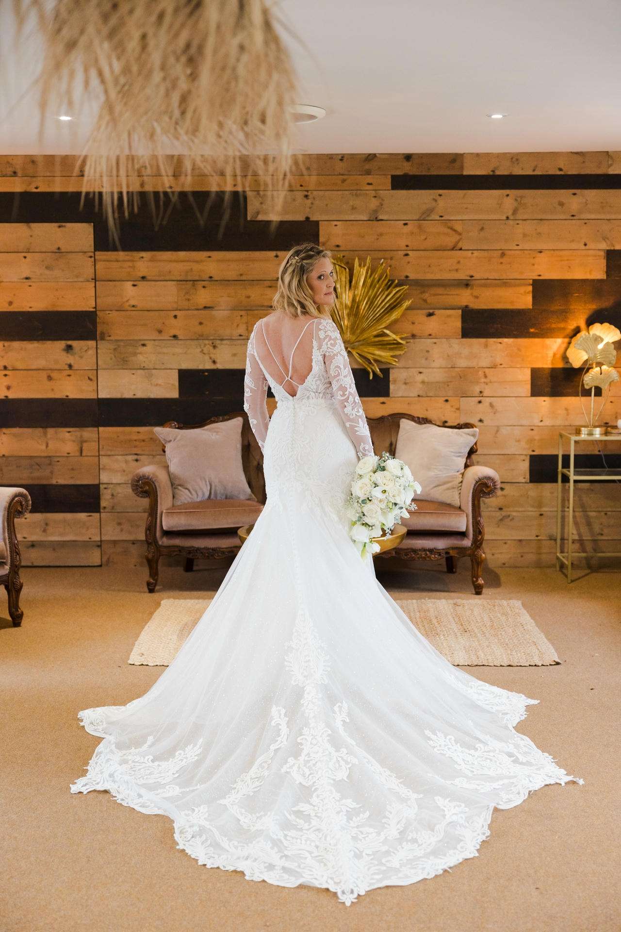 Bride in a white lace wedding gown with a long train and crisscross back stands with a bouquet in a warm wood-paneled room.