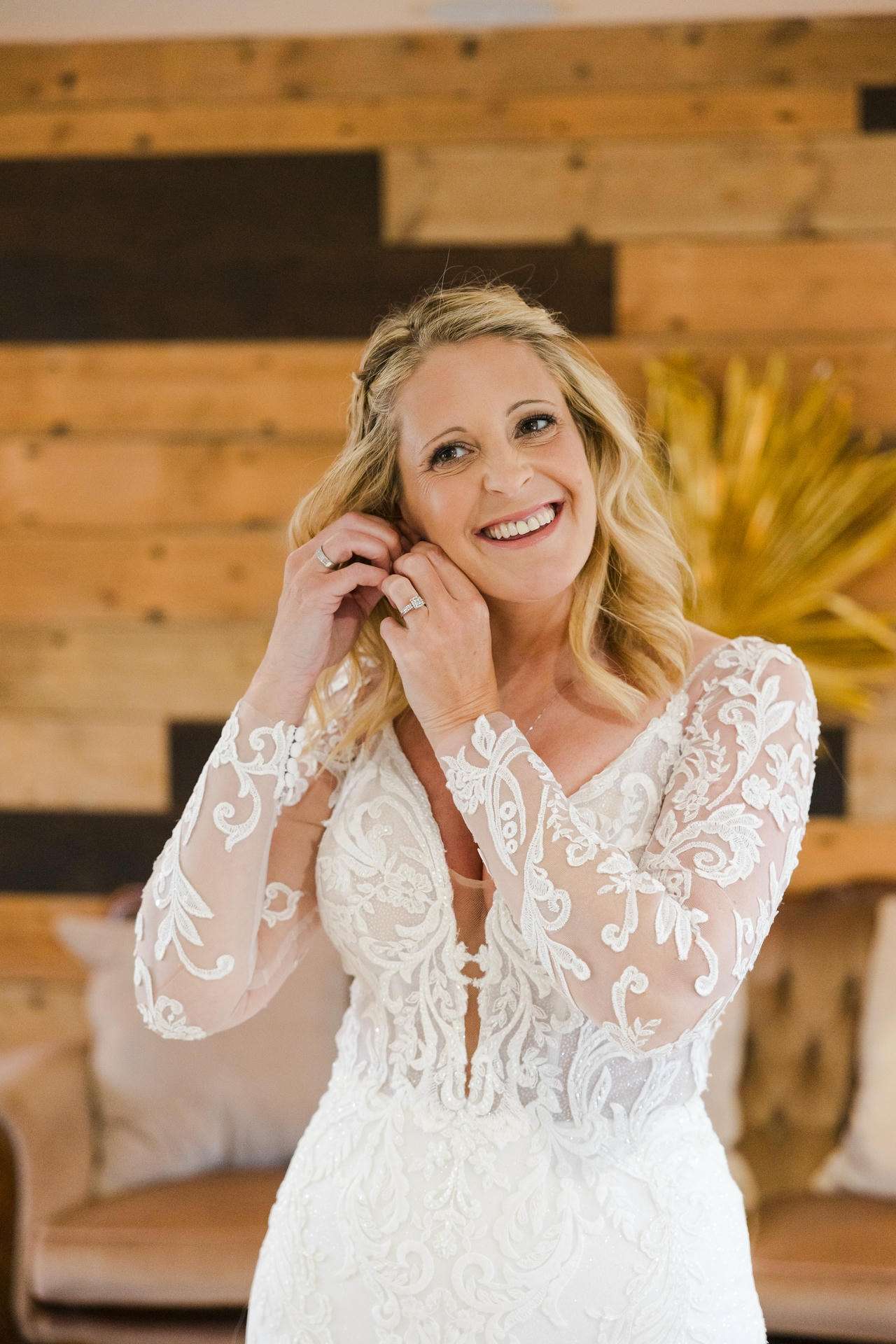 Smiling bride in a white lace wedding gown adjusts her earrings indoors.