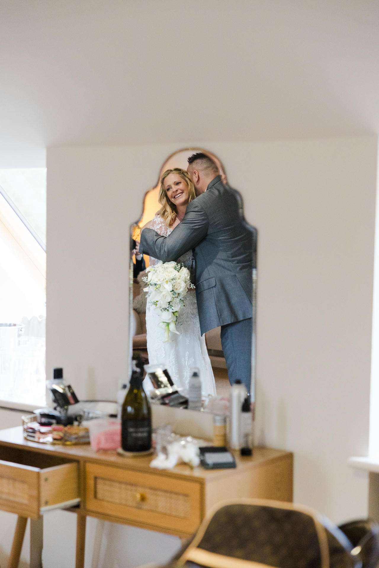 Bride in a white wedding dress holds a bouquet as she embraces her groom in a gray suit, reflected in a dresser mirror.
