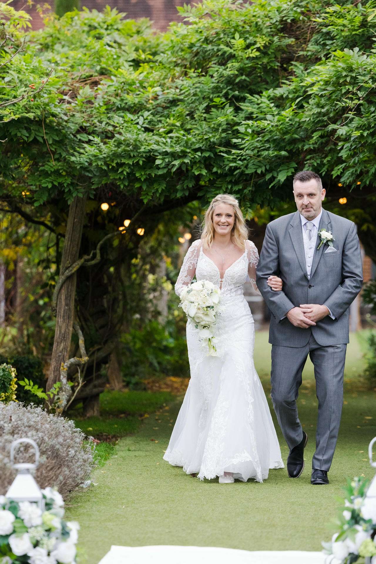 A bride in a white lace gown walks arm-in-arm with a man in a gray suit along a lush garden aisle.