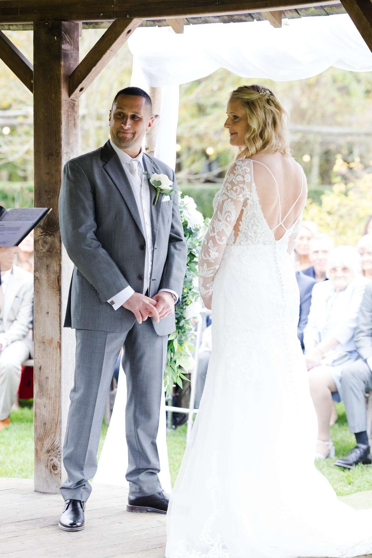 Bride in a white lace gown and groom in a gray suit stand facing each other during an outdoor wedding under a wooden arch.