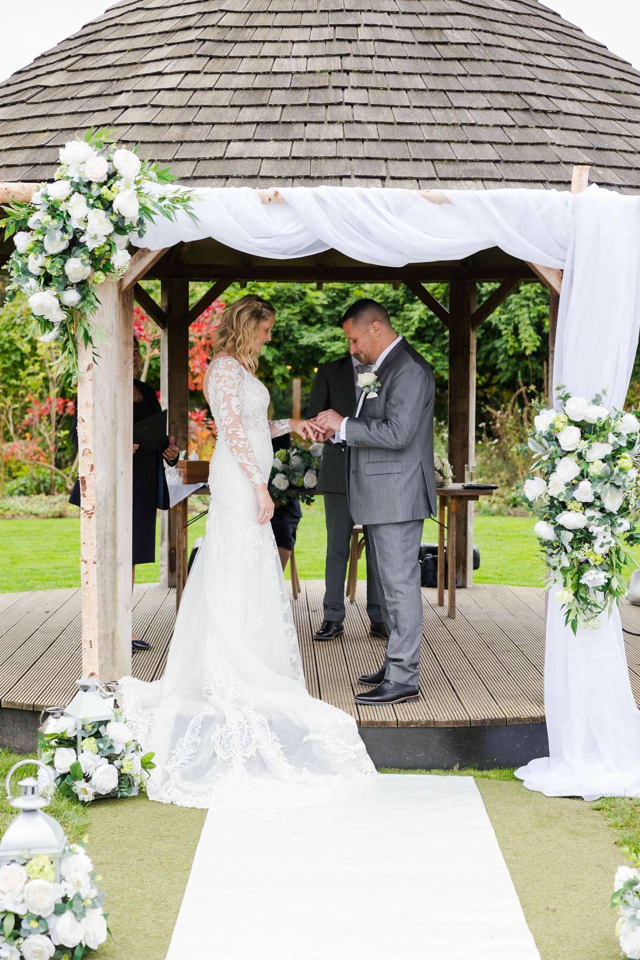 Bride and groom exchange rings under a flower-decorated gazebo during an outdoor wedding.