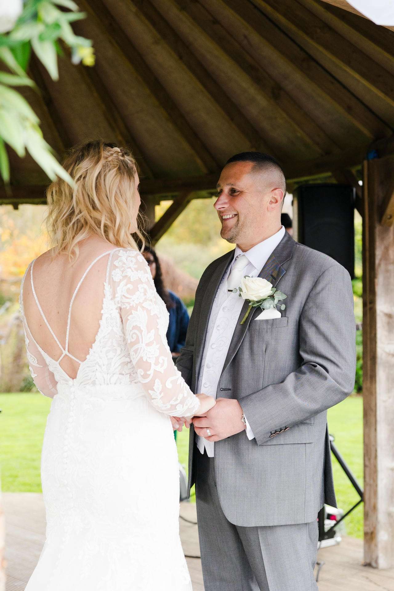 Bride in a lace-back wedding dress and groom in a gray suit hold hands and smile at each other under a wooden pavilion.