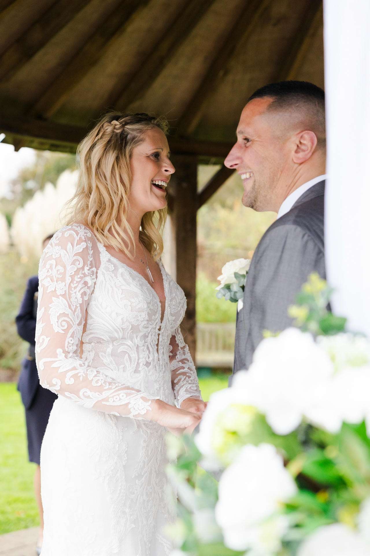 A bride in a white lace gown and a groom in a gray suit smile at each other while holding hands under a wooden gazebo.