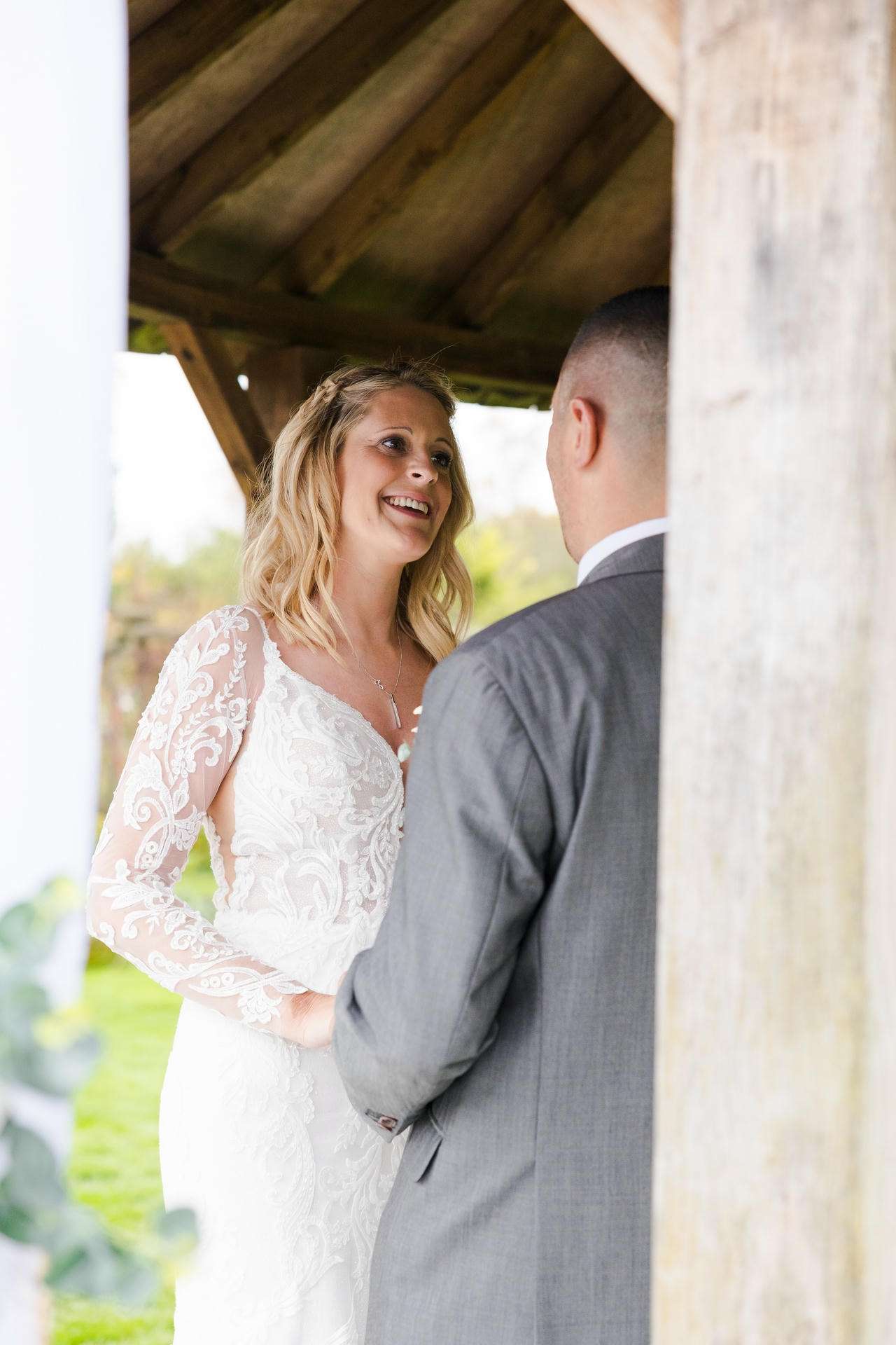 Bride in a lace wedding dress smiles at her groom in a gray suit under a wooden shelter.