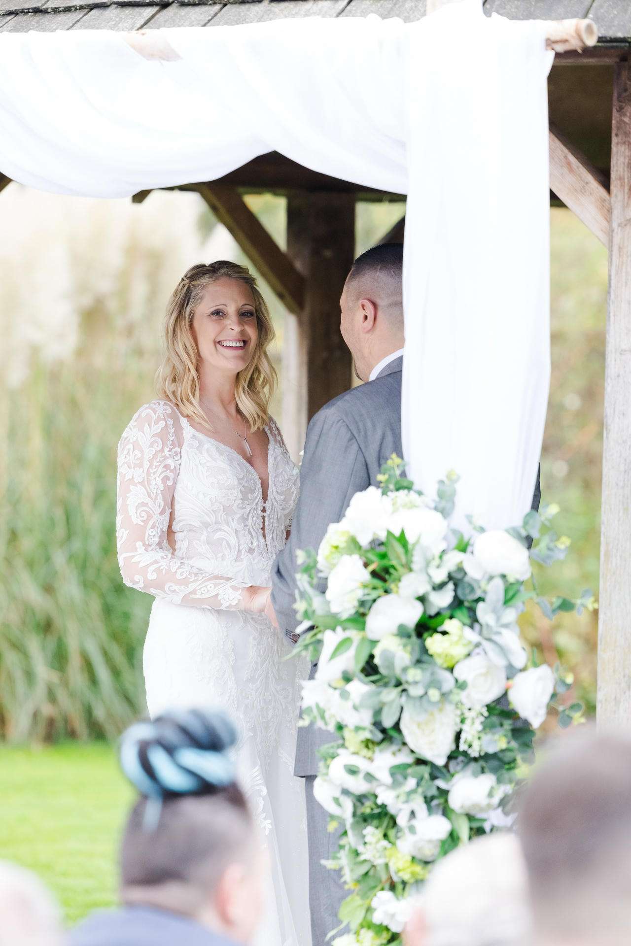Bride and groom stand facing each other under a white fabric arch at an outdoor wedding, with a large white and green bouquet in the foreground.