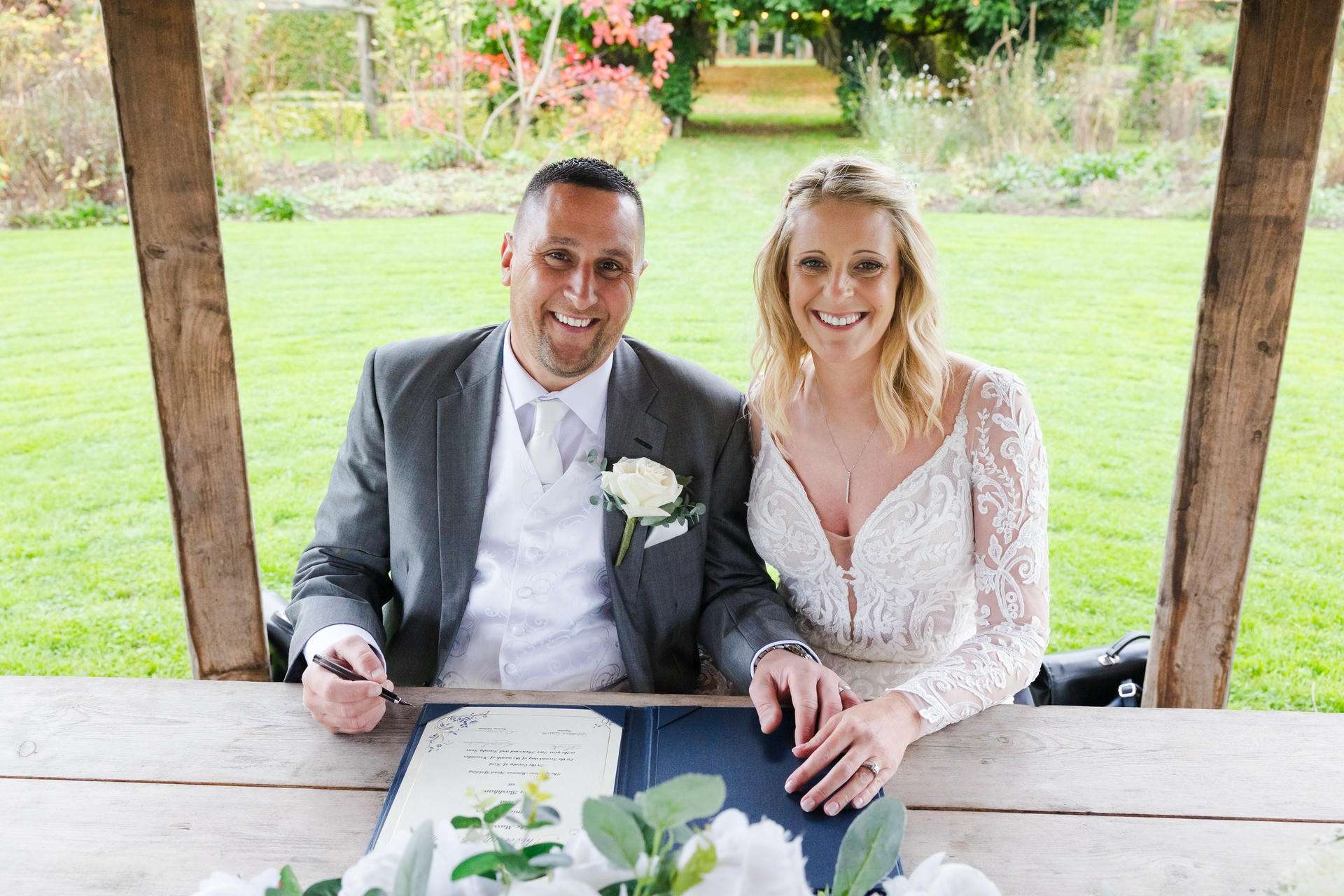 Smiling bride and groom sign a document at an outdoor wedding under a wooden pergola.