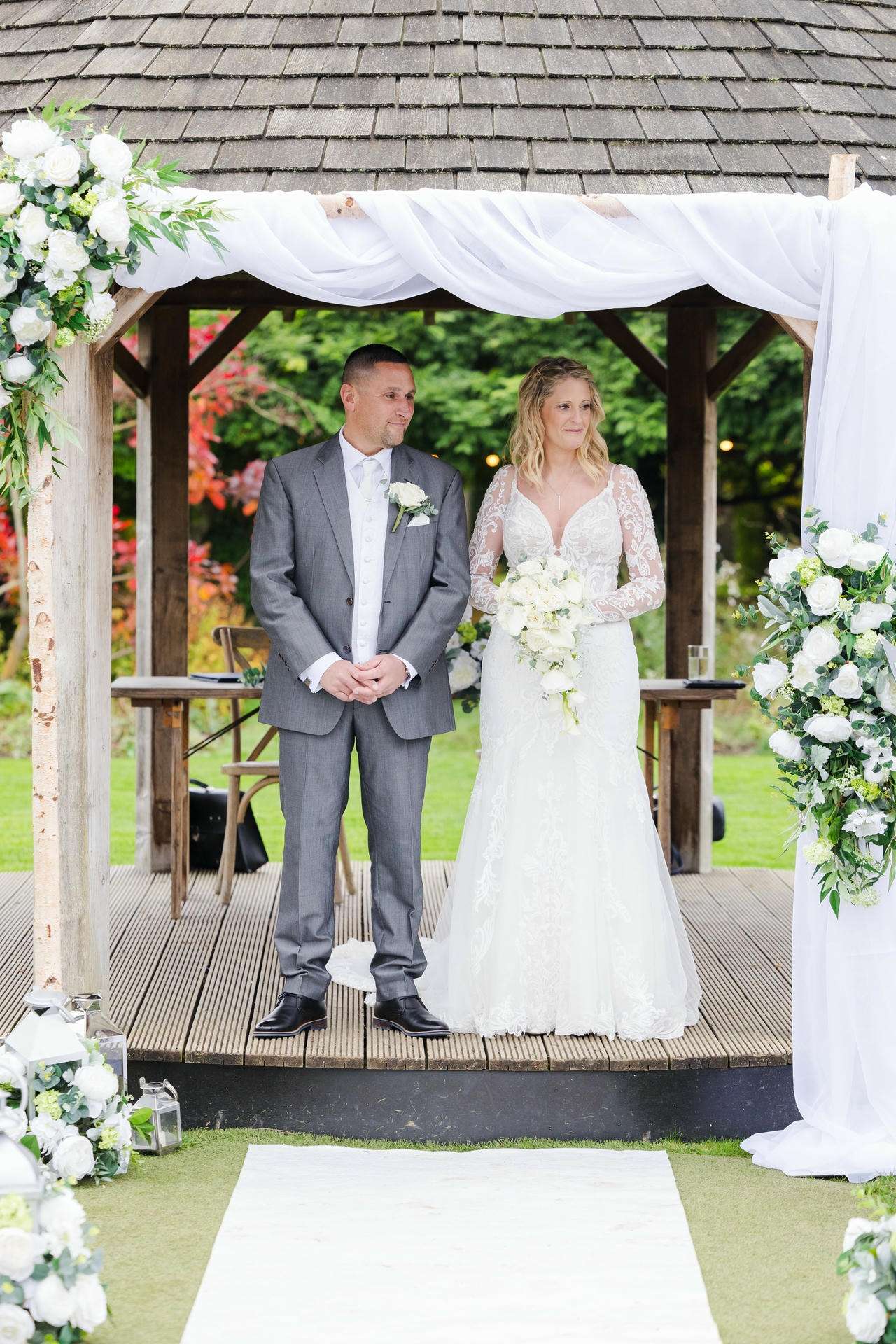 Bride in a lace white wedding gown holds a bouquet as she stands with the groom in a gray suit under a white-draped wooden arch adorned with flowers.