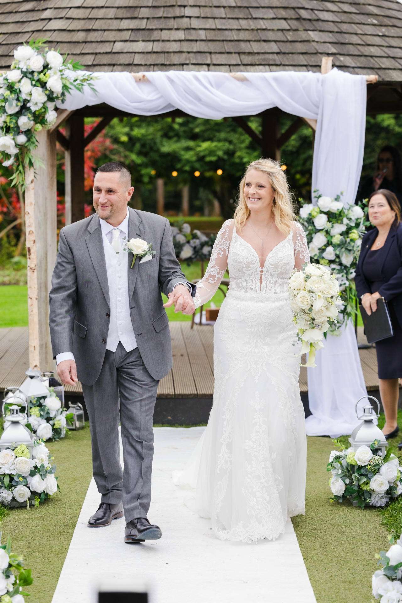 Bride in a lace wedding dress carrying a white bouquet walks beside the groom in a gray suit under a white-draped floral arch at an outdoor wedding.